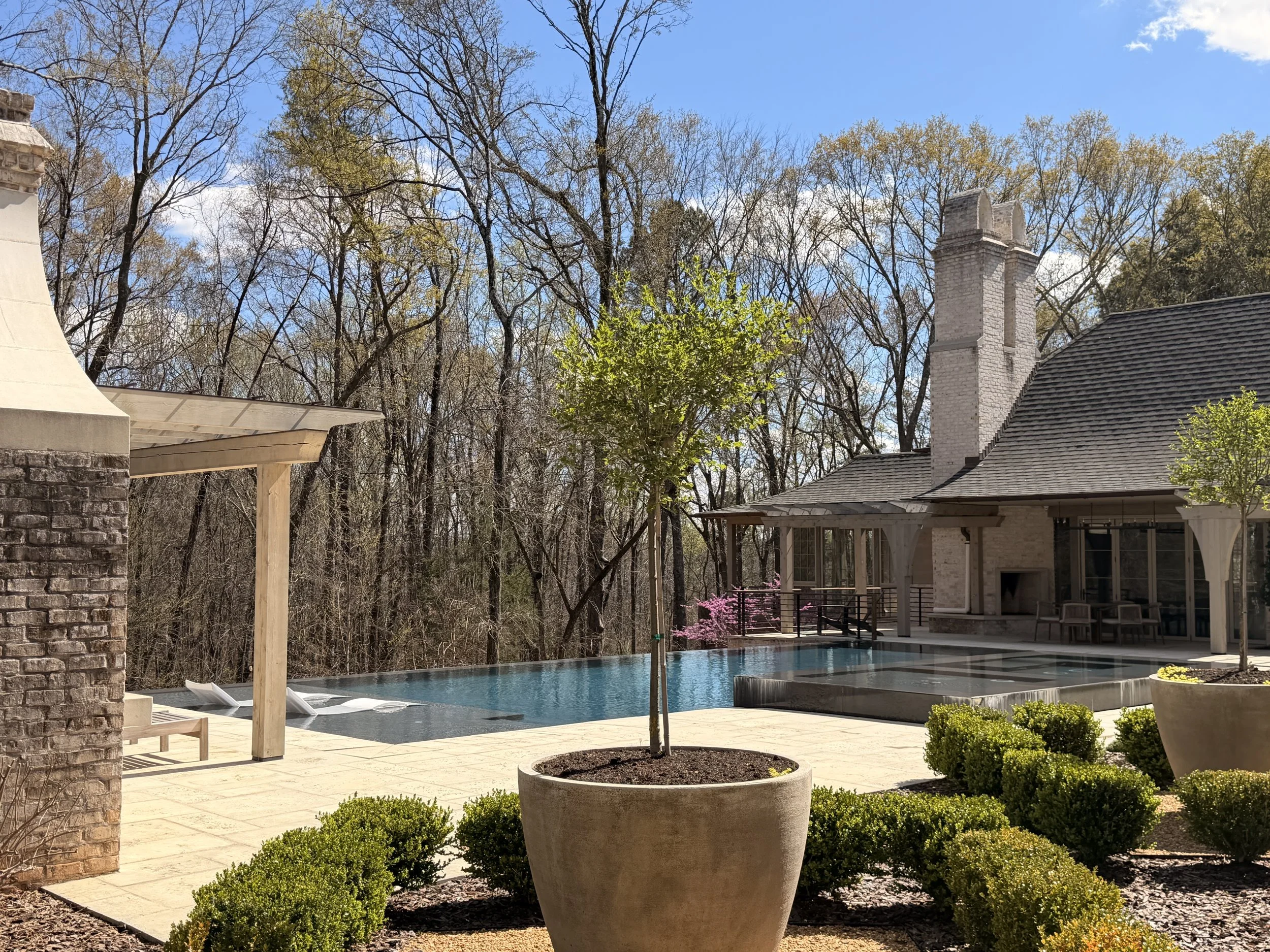 Residential backyard with a swimming pool, surrounded by potted plants and small shrubs, with a house featuring a chimney and a patio, and trees in the background on a clear, sunny day.