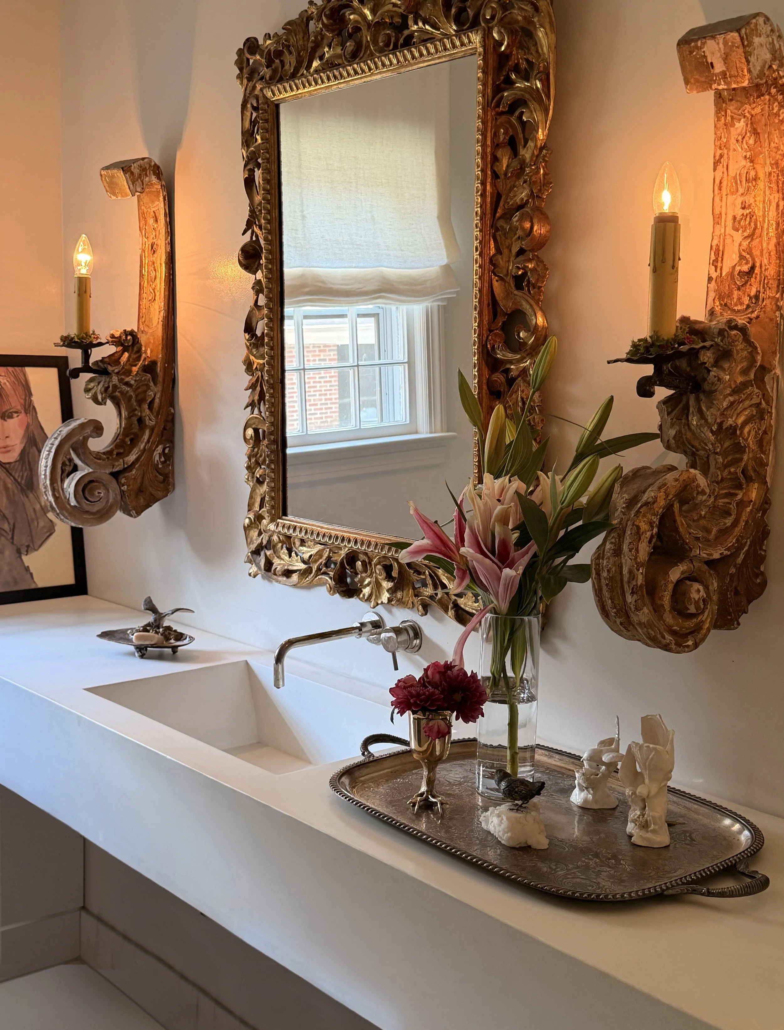 A decorative white kitchen sink unit with a silver tray holding various animal sculptures, including a carved bird and abstract shapes. A vase with lilies and roses is on the right, and a large ornate gold-framed mirror hangs above, reflecting a wind