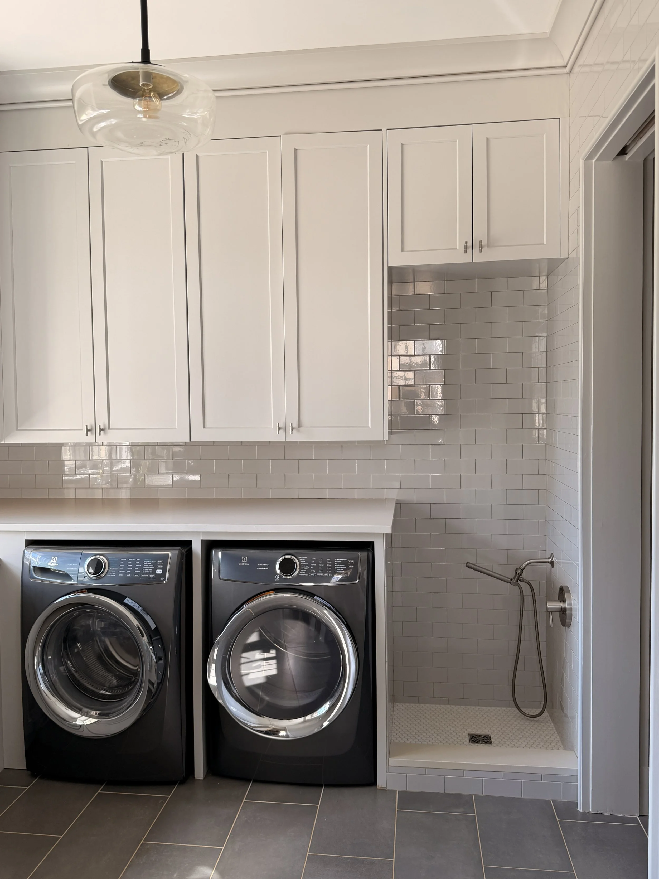 A laundry room with white cabinets, a black front-loading washer and dryer, and a shower area with beige tile walls and a handheld shower head.