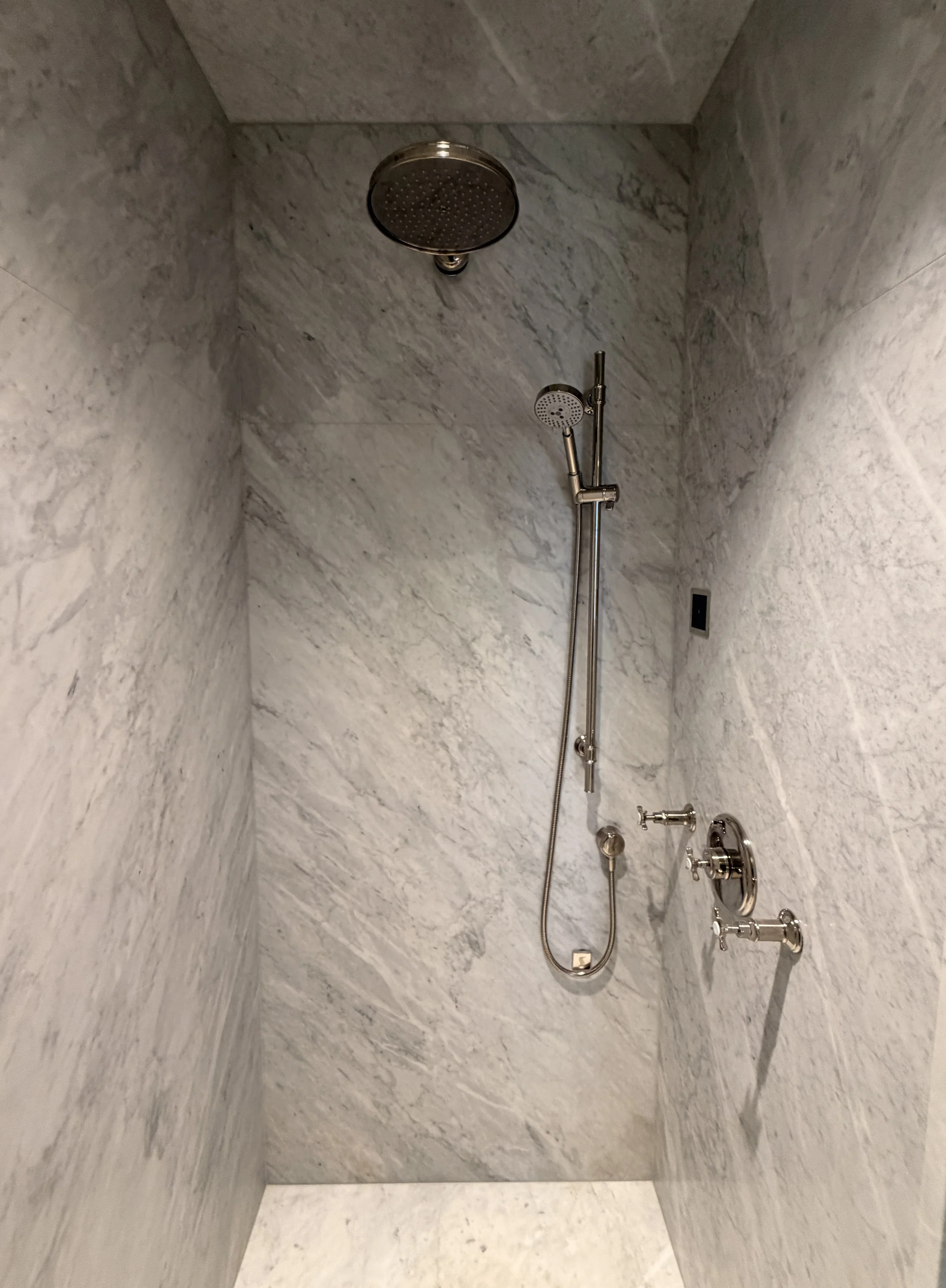 A walk-in shower with marble walls, a rainfall showerhead on the ceiling, a handheld showerhead, and a control panel with multiple knobs.