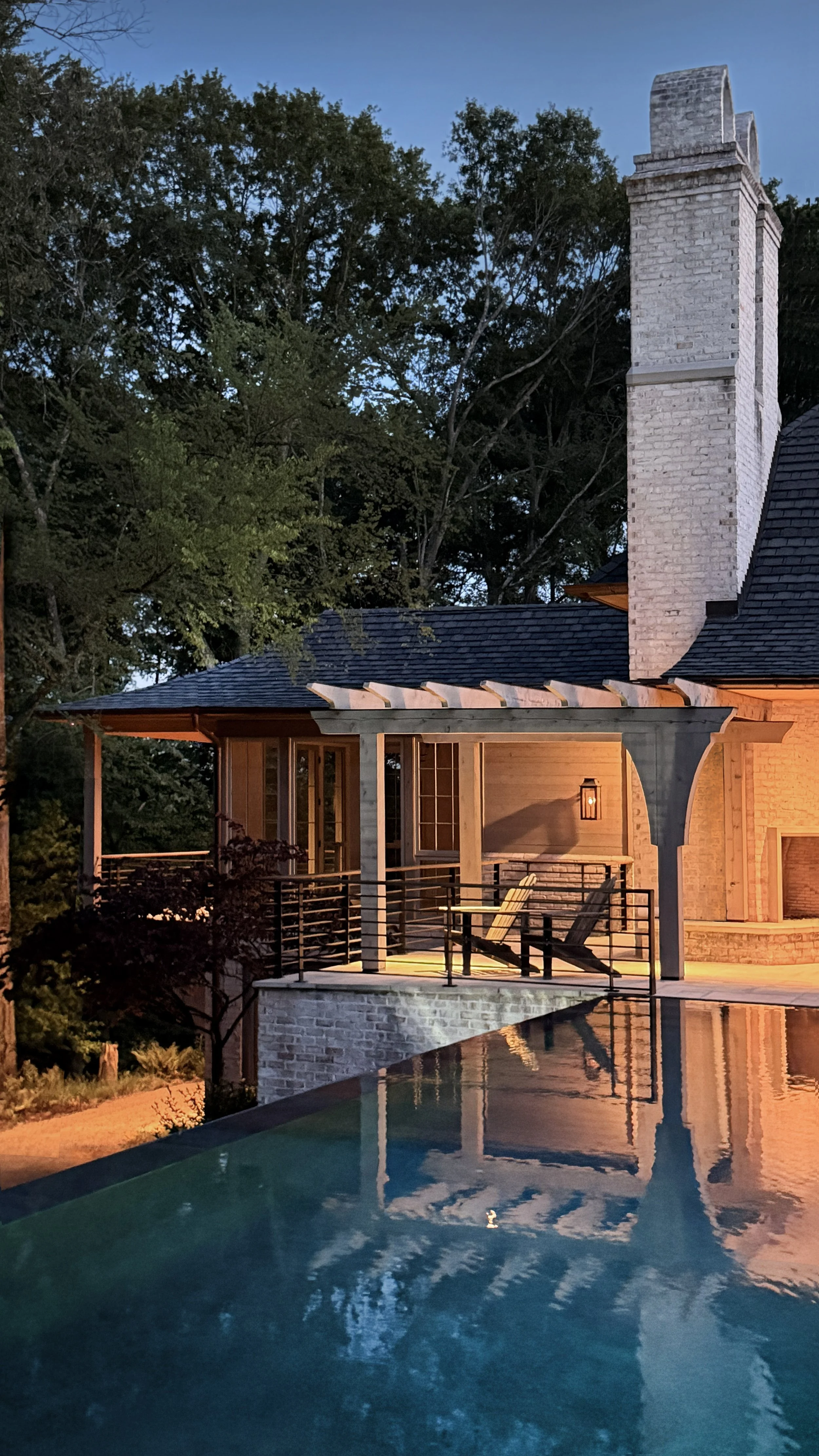 A house with a tall brick chimney, a wooden porch with chairs, a nearby swimming pool, and trees in the background during evening.