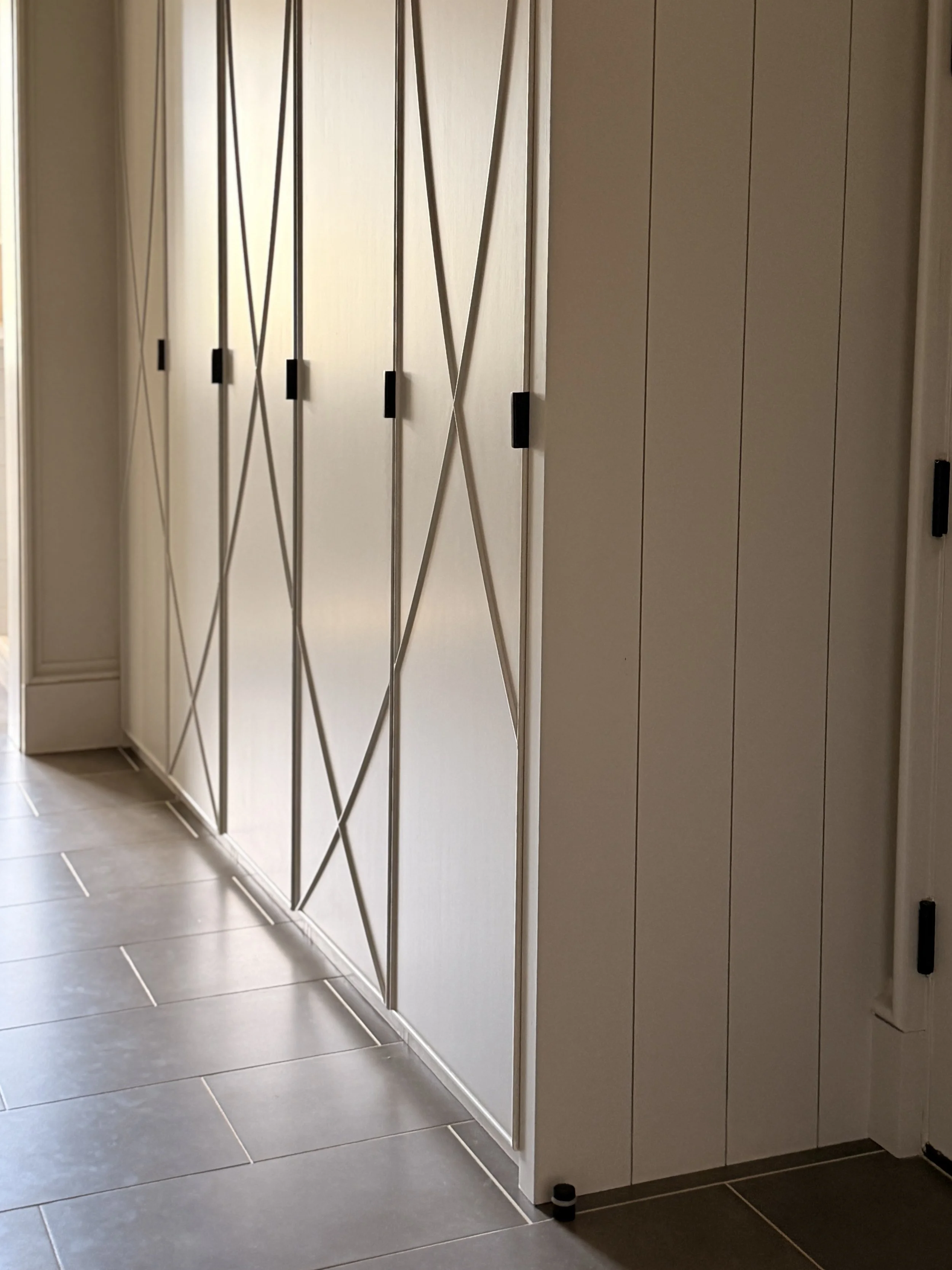 A set of modern, cream-colored closet or cabinet doors with black handles and a decorative metal grid on the left side. The doors are situated in a room with grey tiled flooring.