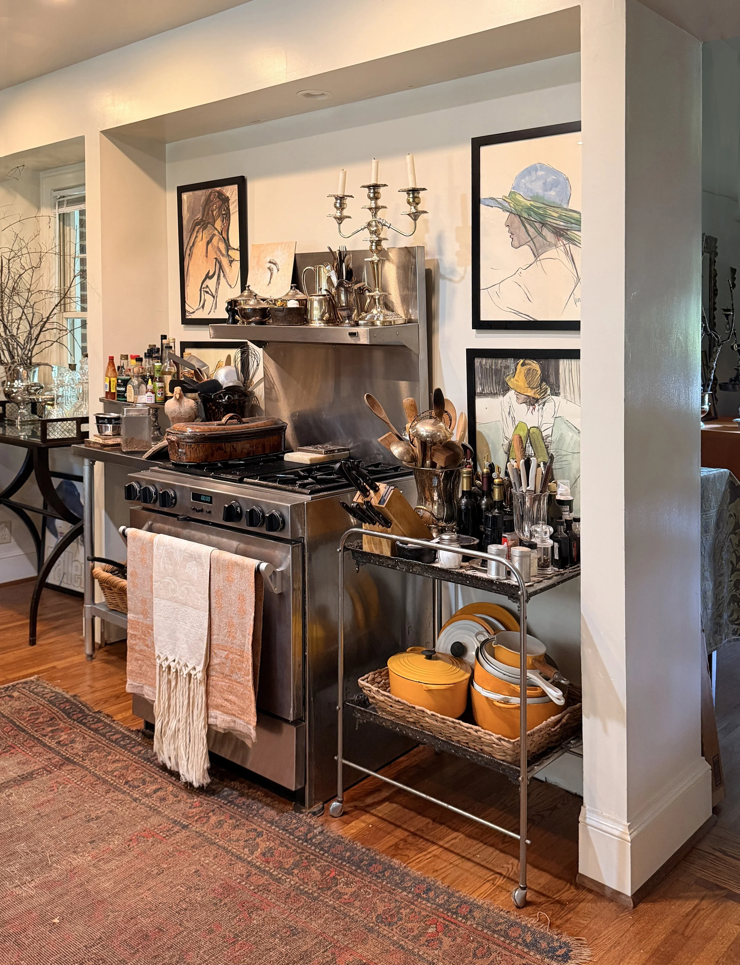 A kitchen wall with three framed line art portraits of women, a stainless steel stove and hood, with cooking utensils and jars on a cart and countertop. Items include yellow bowls, a basket, metal cookware, and various condiments.