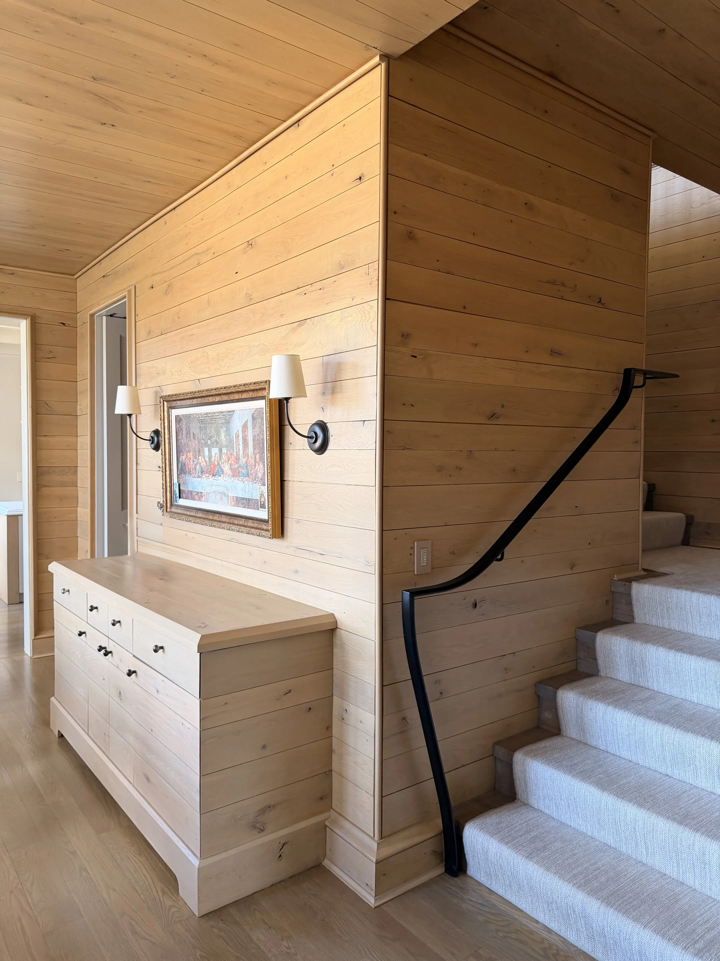 Interior of a home with wood-paneled walls and ceiling, a white dresser with multiple drawers, framed artwork on the wall, two wall-mounted lamps, and a staircase with beige carpeted steps and a black handrail.