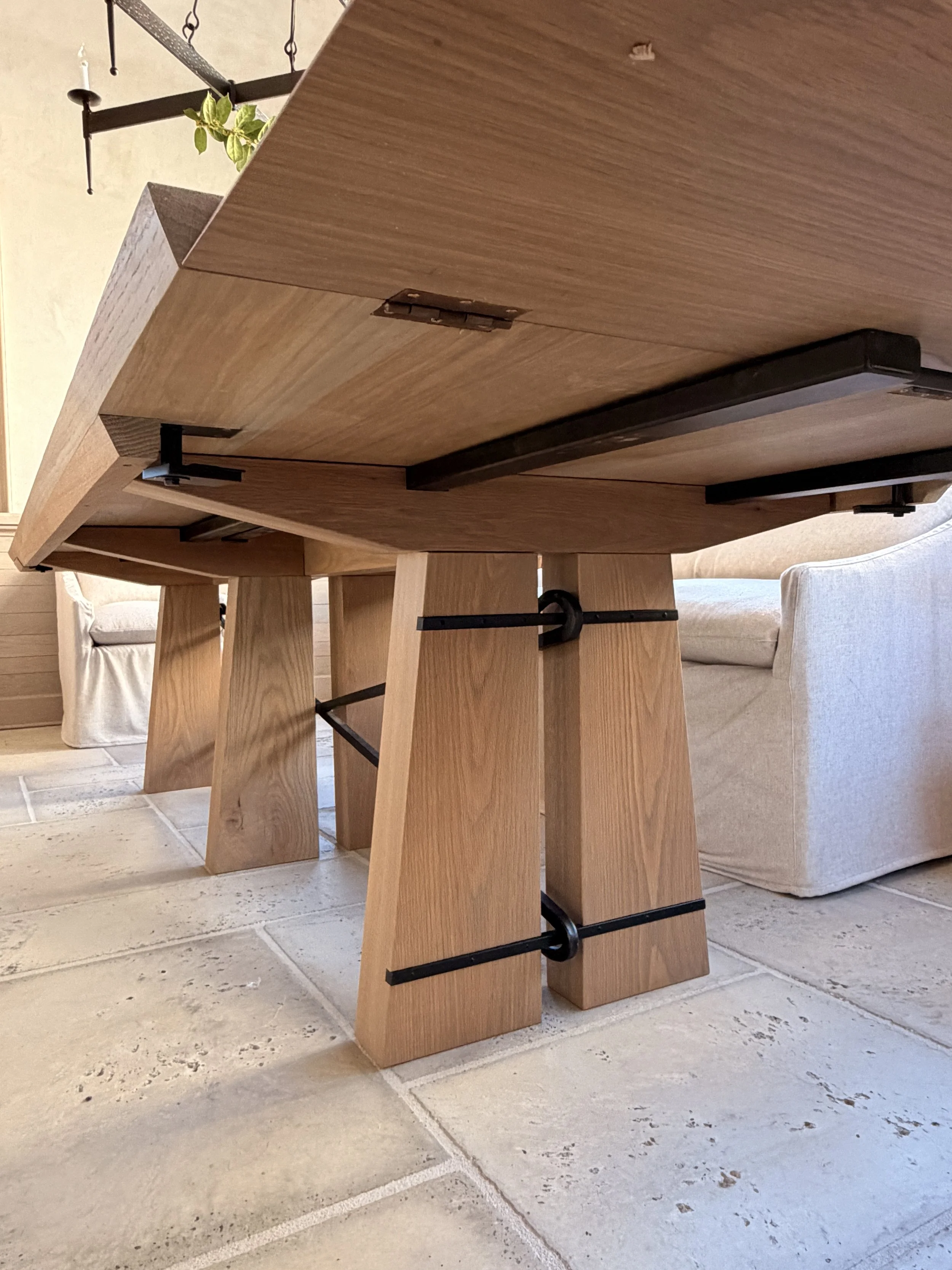 Close-up of a wooden dining table with black metal support beams, positioned on a tiled floor. Part of a beige cushioned chair and a sofa are visible in the background.