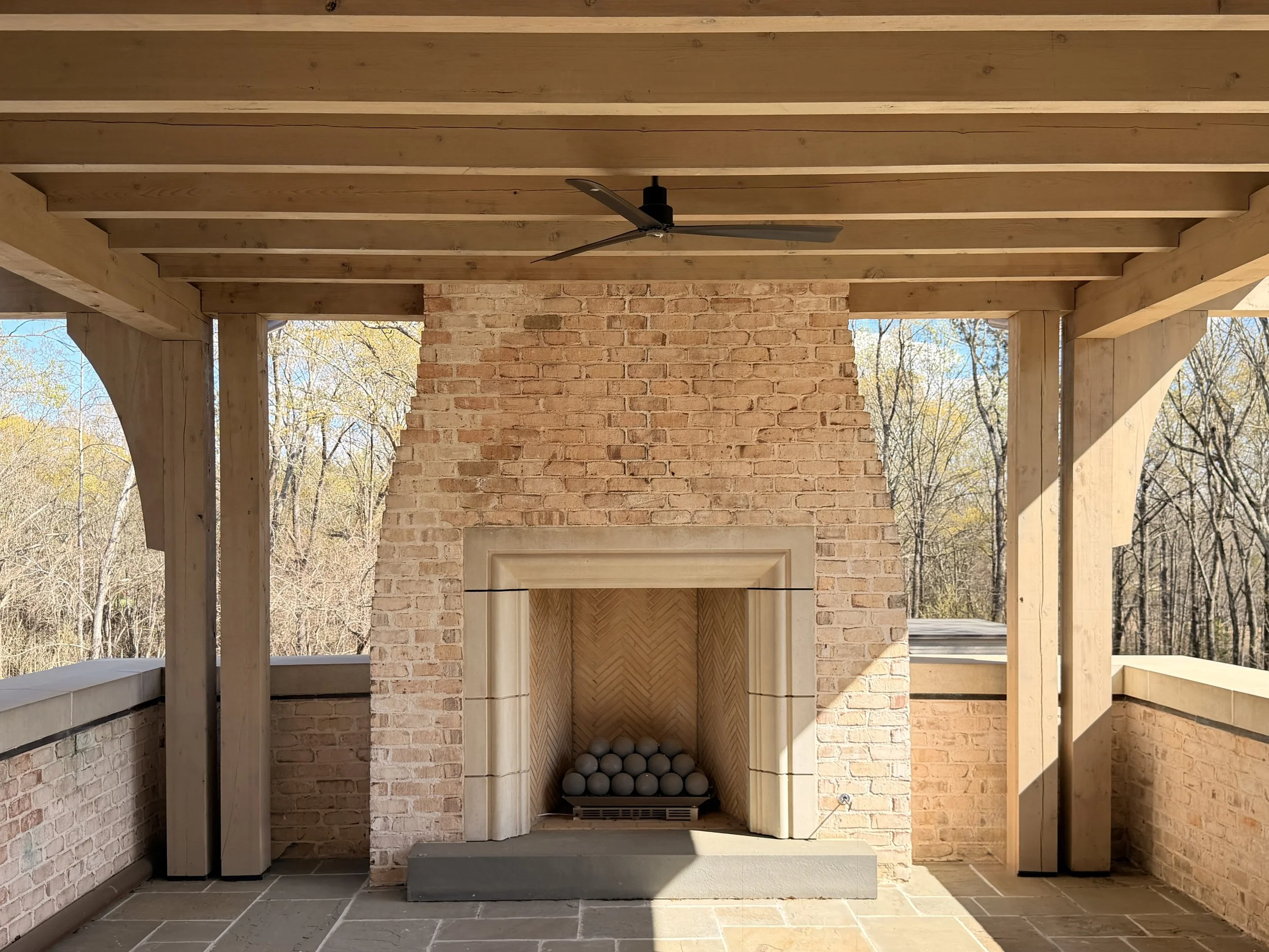 The image shows a construction area with exposed wooden beams on the ceiling, a brick chimney as a focal point, and a firepit with balls inside. There are trees visible in the background through open spaces, indicating an outdoor setting.