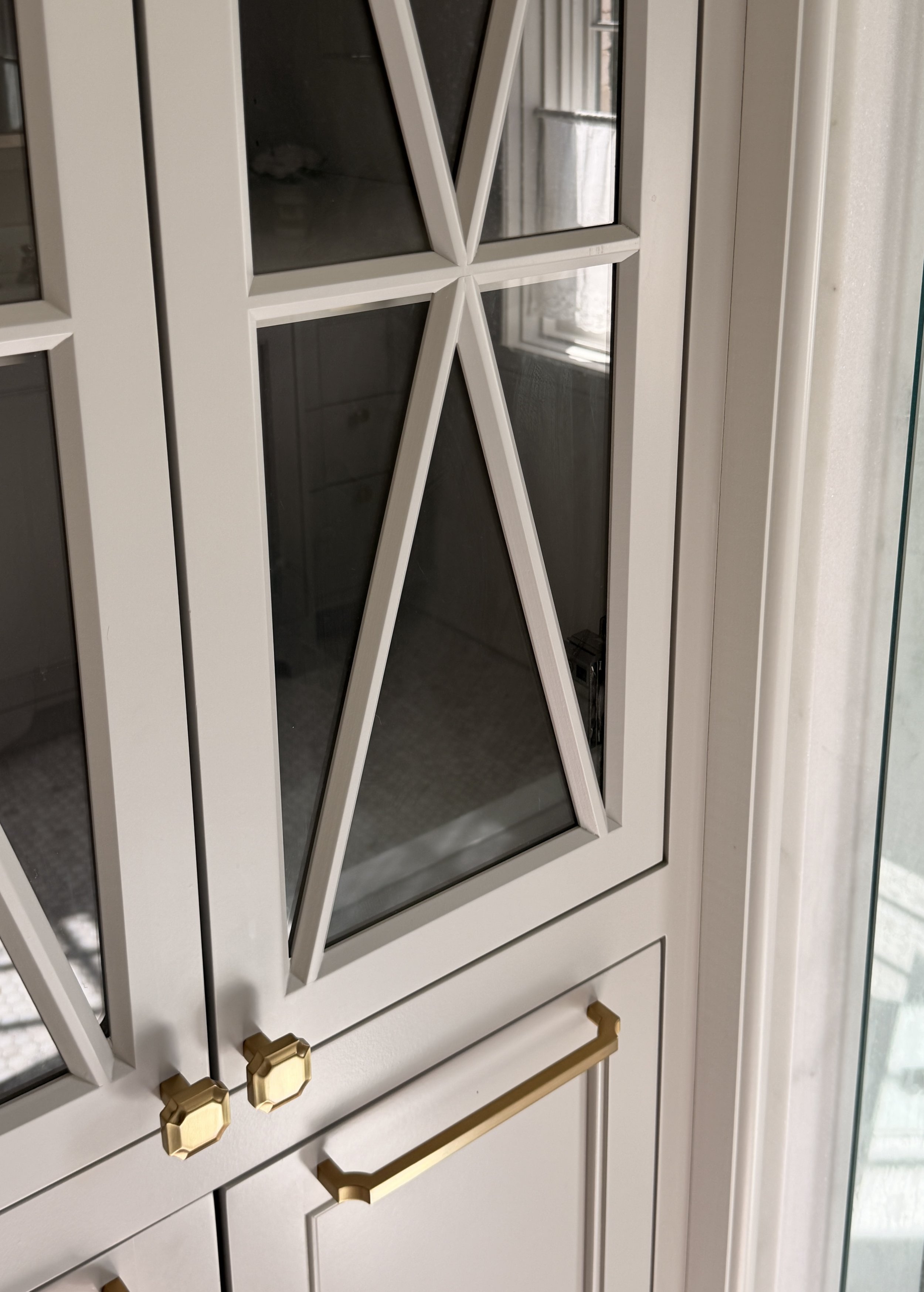 A white cabinet with glass doors and gold hardware, including gold drawer pulls and a gold handle, photographed indoors with natural light.
