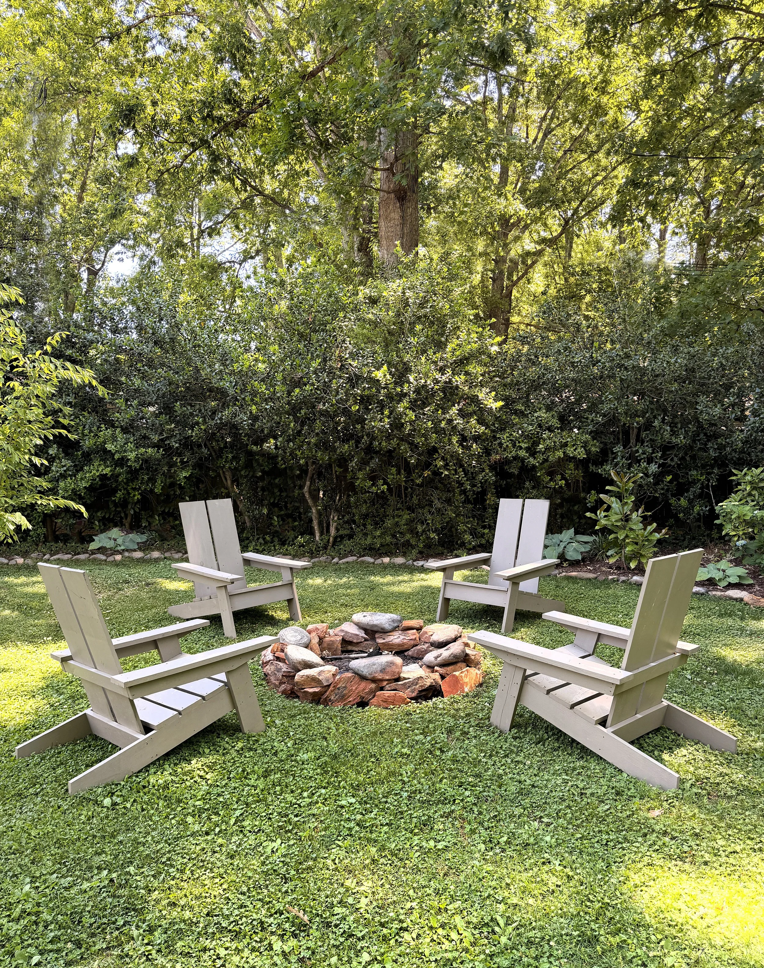 A backyard with a circle fire pit made of bricks surrounded by six white Adirondack chairs, with green grass, trees, and shrubs in the background.