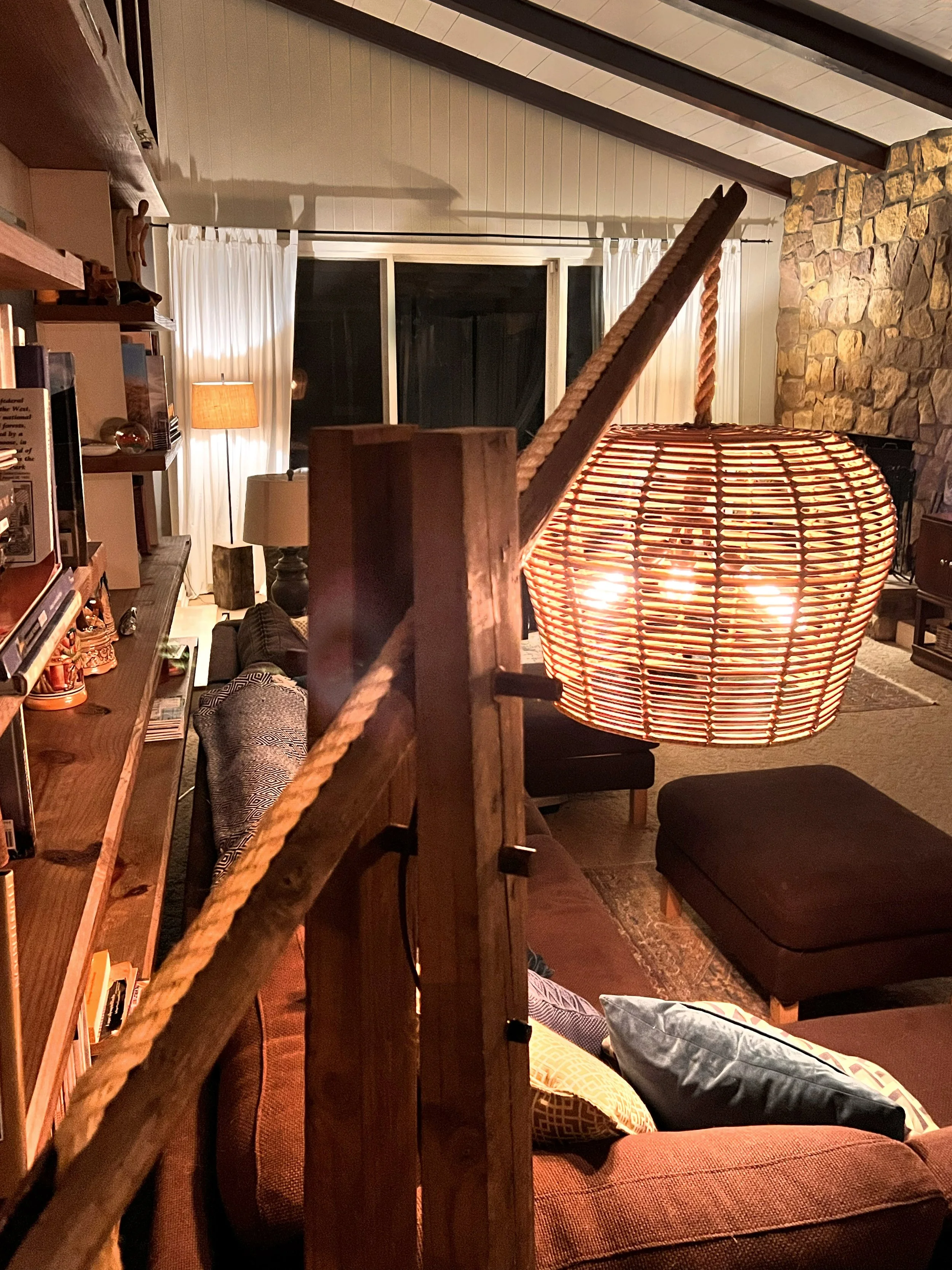 View of a cozy living room at night with a lit paper lantern hanging in the foreground, a couch with pillows, a stone wall, a window with curtains, and various lamps illuminating the space.
