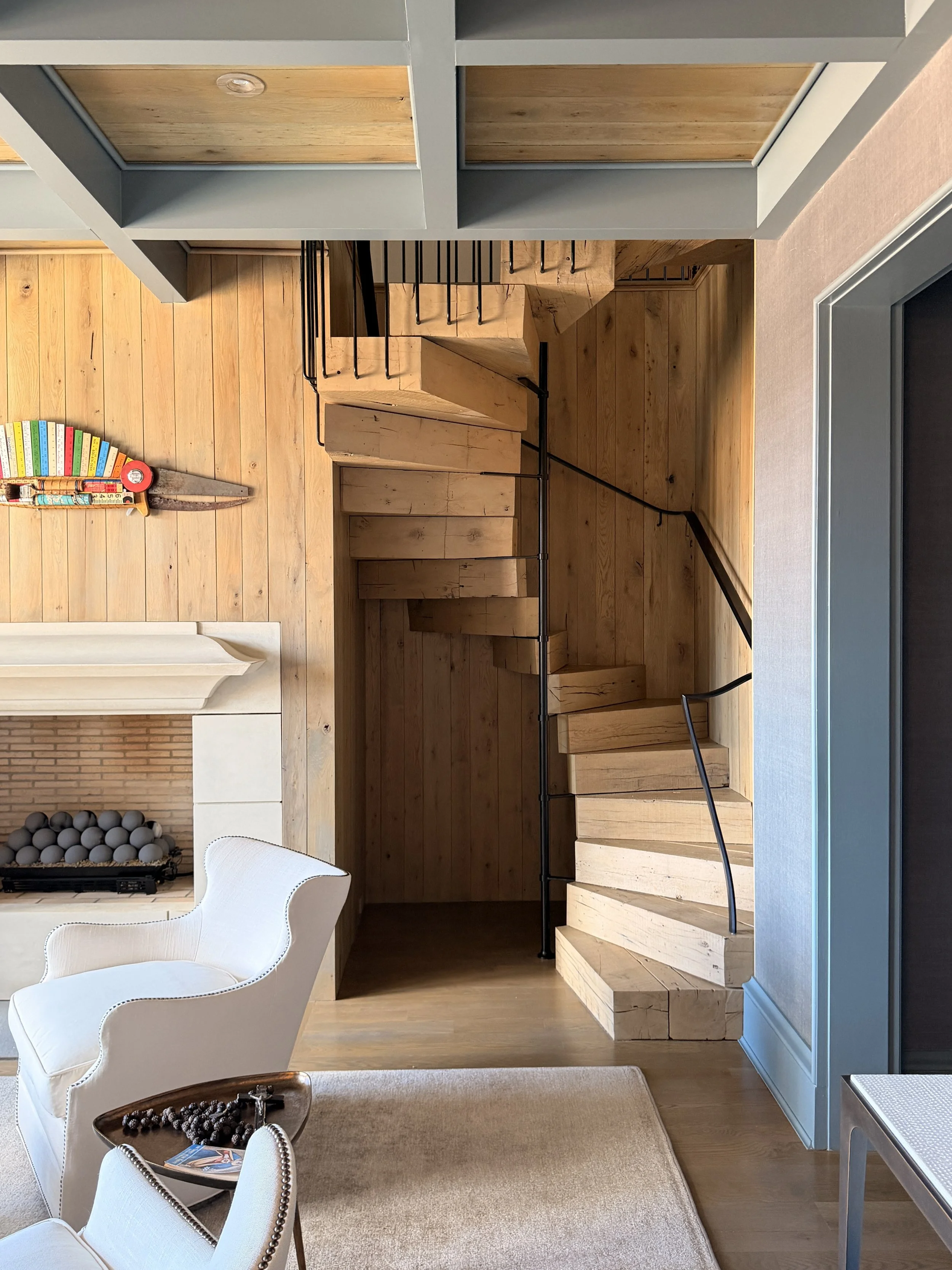 Interior view of a room with wooden flooring, white armchairs, a coffee table with pearls and a magazine, a beige carpet, and a staircase with wooden steps and black metal railing. There is a fireplace with black stones and a vent, and colorful books