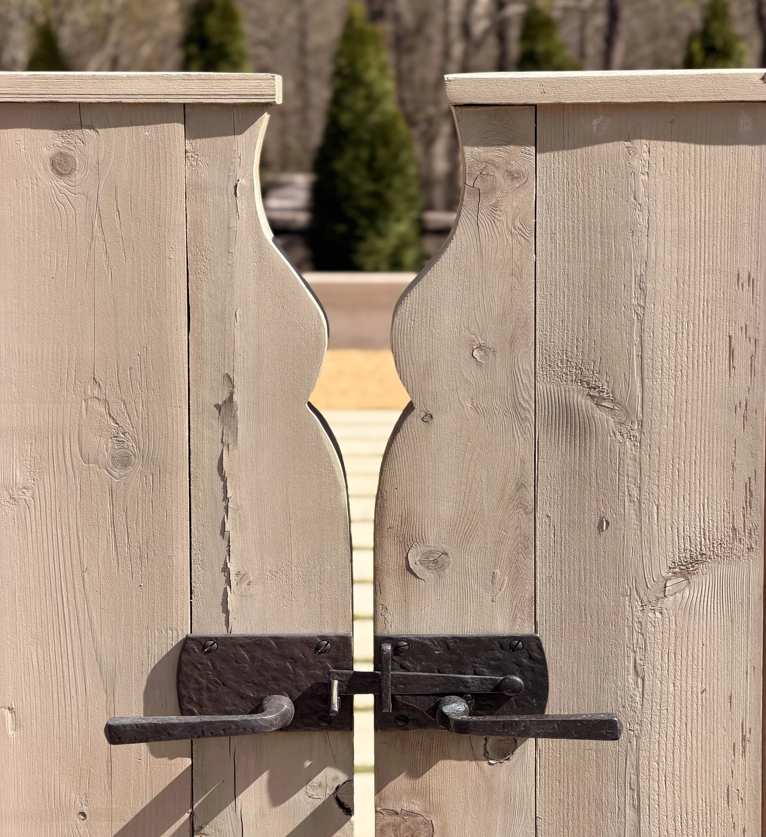 Close-up of a white wooden gate with black hinges, partially open, showing trees with blurred foliage in the background.