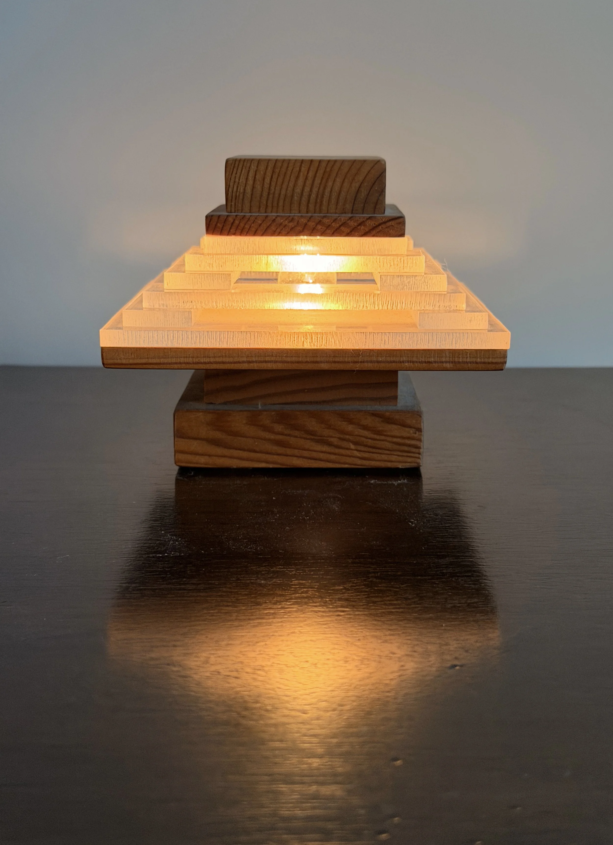 A wooden pyramid-shaped lamp with multiple layers illuminated, placed on a dark surface, casting a warm glow and reflected on the surface below.