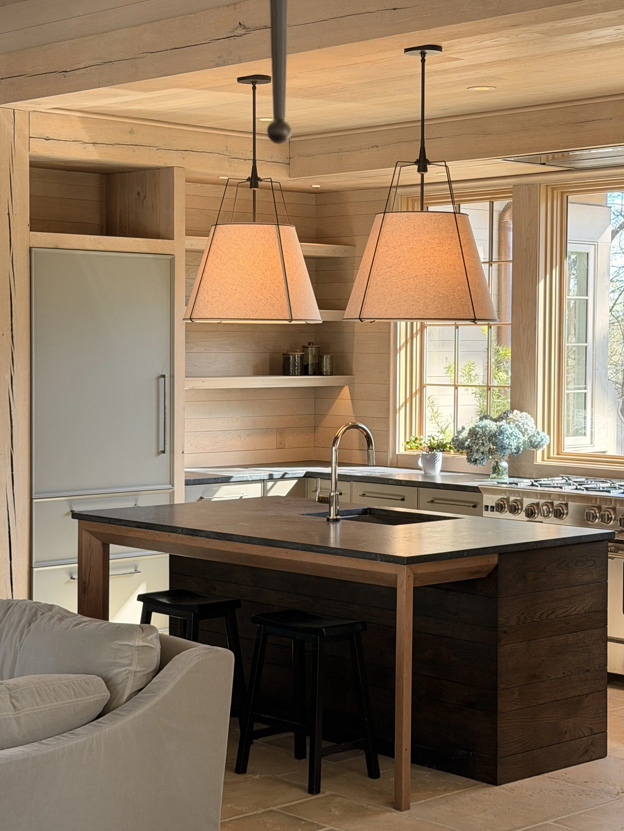 A kitchen with wooden cabinets and walls, a black granite countertop, a white sink with a chrome faucet, two beige pendant lights hanging above a wooden kitchen island, a large window with a flower arrangement, and a beige sofa partially visible in t