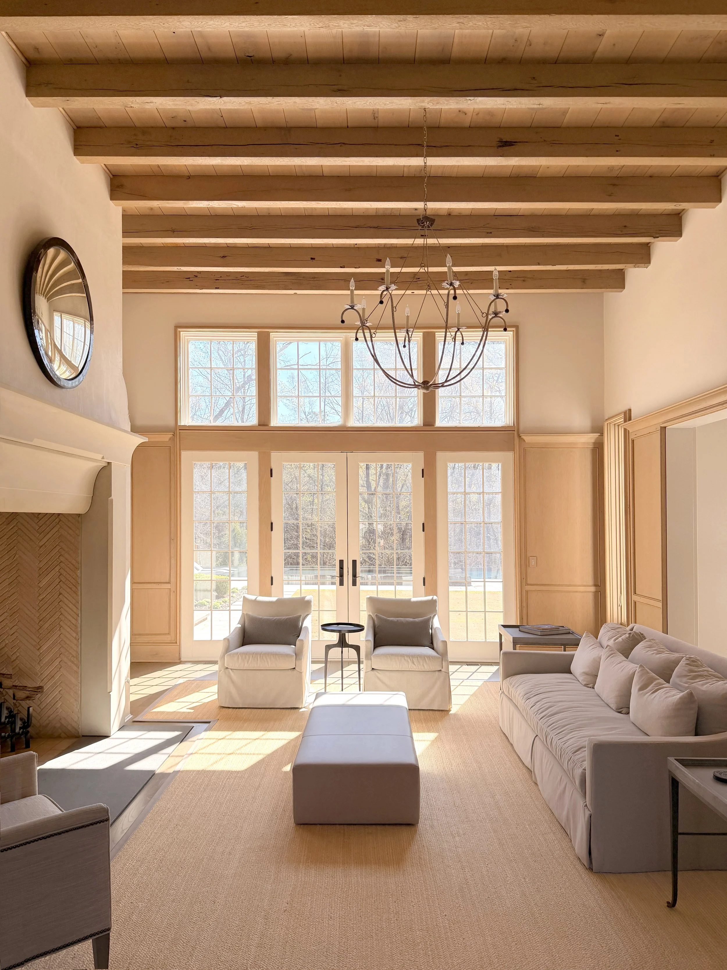 Bright living room with large windows, a white sofa, two matching armchairs, a small round table, and a chandelier hanging from the wooden ceiling beams.