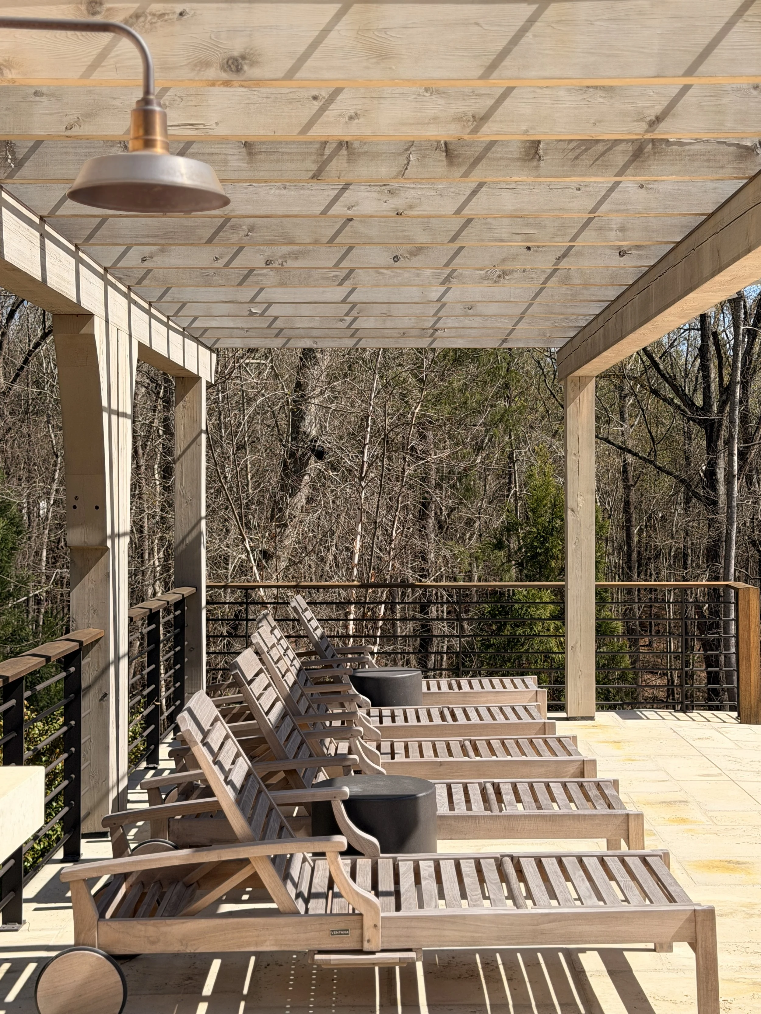 Outdoor patio with wooden furniture and a shaded pergola, overlooking trees and a railing.
