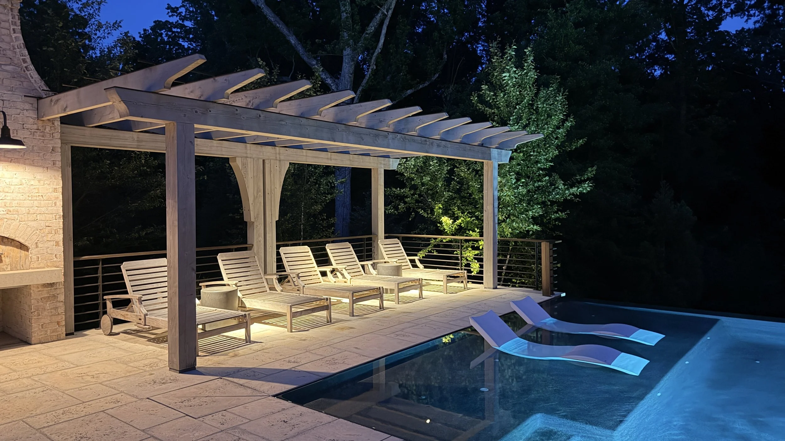Nighttime outdoor scene featuring a wooden pergola with lounge chairs underneath, adjacent to a swimming pool with floating lounge chairs, surrounded by lush trees and foliage.