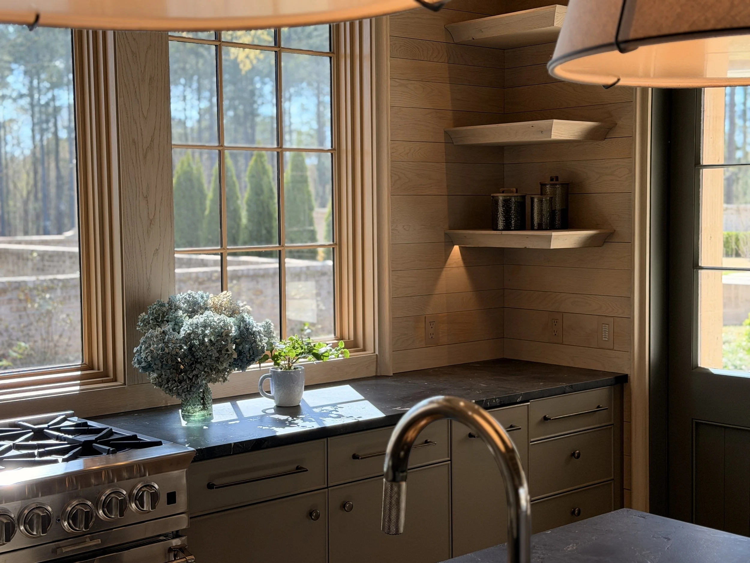 A kitchen with large window, wooden walls, flowers in a vase, and a stove.