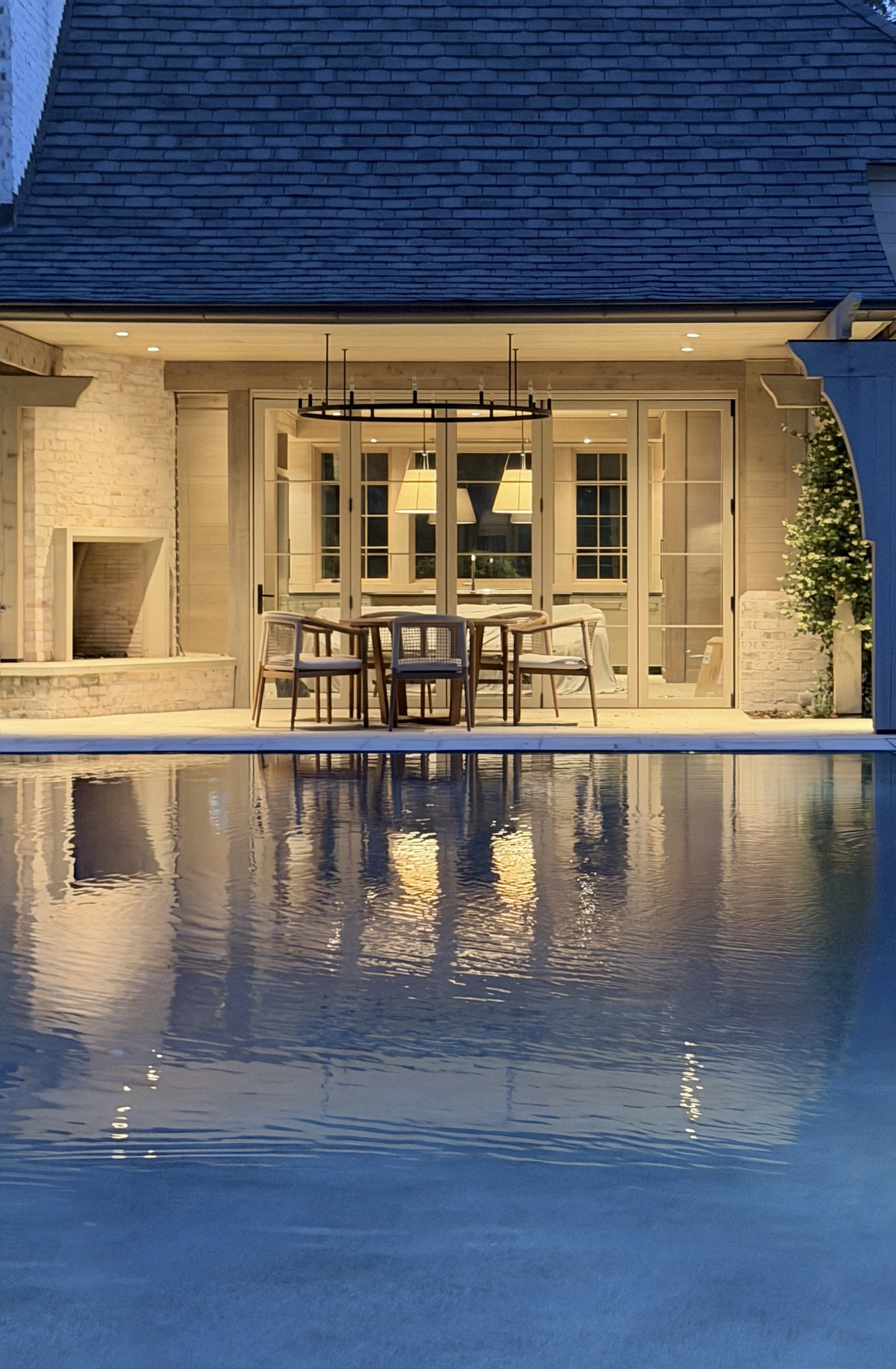 A house with a brick and timber exterior, glass door, and a wooden table with chairs on a patio, reflected in a swimming pool at dusk.