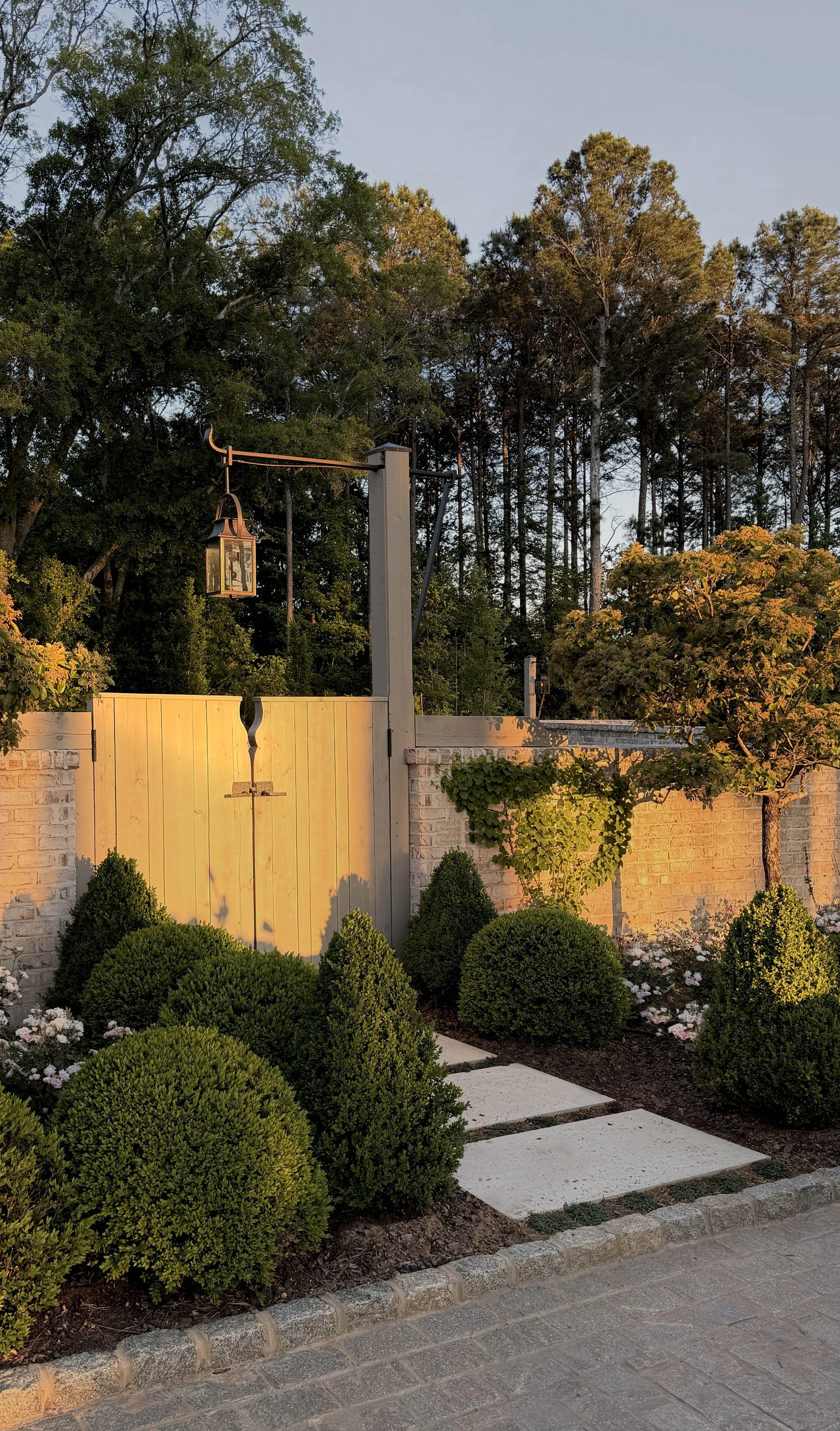 A garden with trimmed bushes, flowers, a paved walkway with stepping stones, a beige wooden gate, a brick wall, and a black lantern-style light fixture hanging from a pole. Tall trees in the background under a clear sky at sunset.