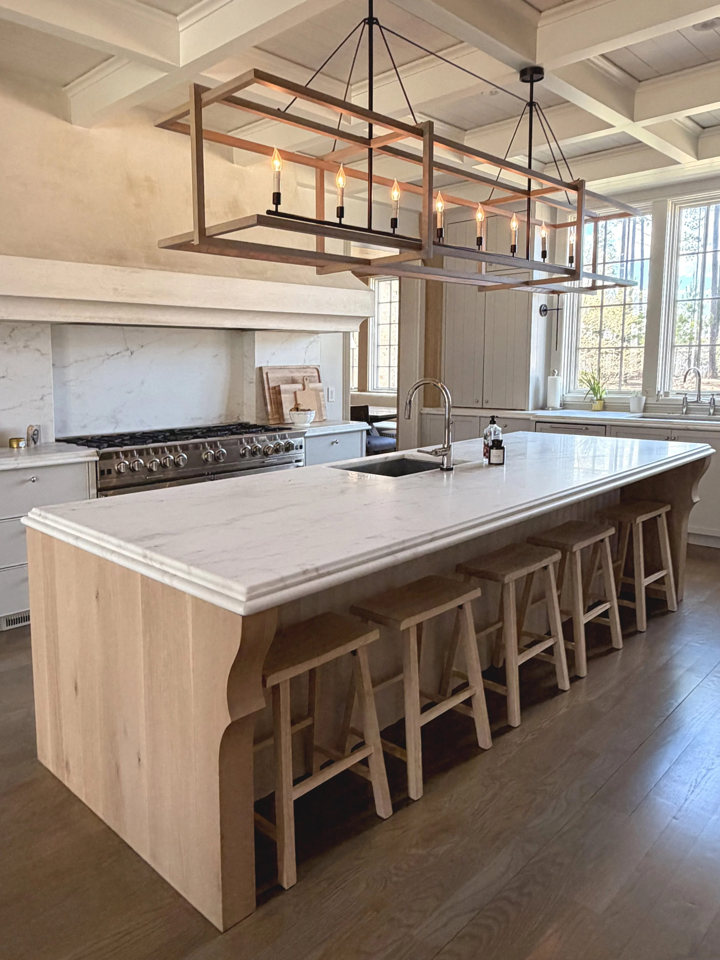 A modern kitchen with a large marble island, wooden bar stools, a chandelier with candle-shaped lights, white cabinets, a marble fireplace, and large windows letting in natural light.