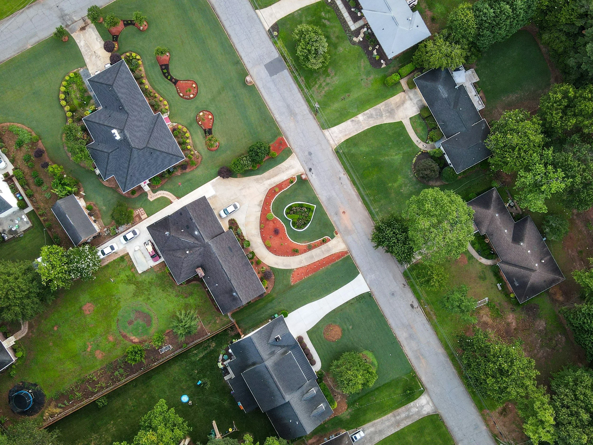 Aerial view of a suburban neighborhood with landscaped yards, houses, greenery, trees, and a paved road.