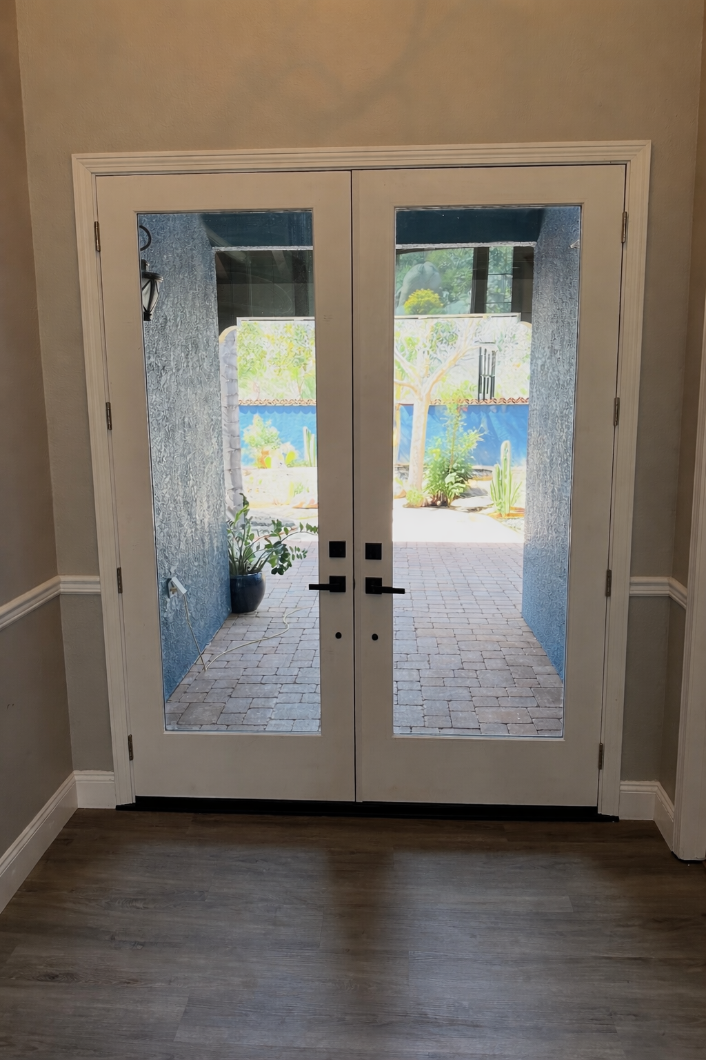 Interior view of glass double doors leading to a backyard with brick patio, cactus, trees, and garden plants.