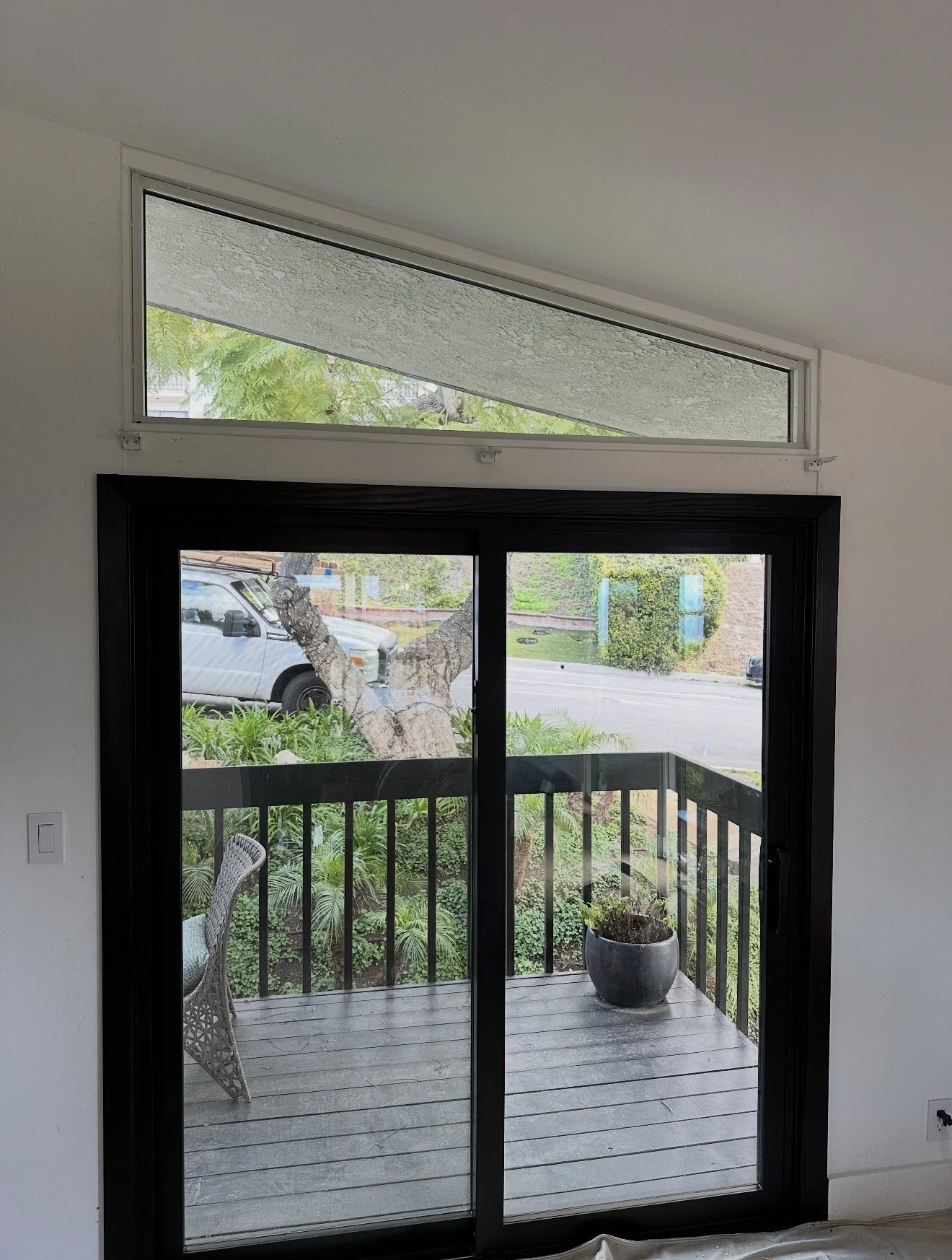 Indoor view of a glass sliding door leading to a small outdoor balcony with a potted plant, balcony railing, and view of a tree and parked vehicle outside.