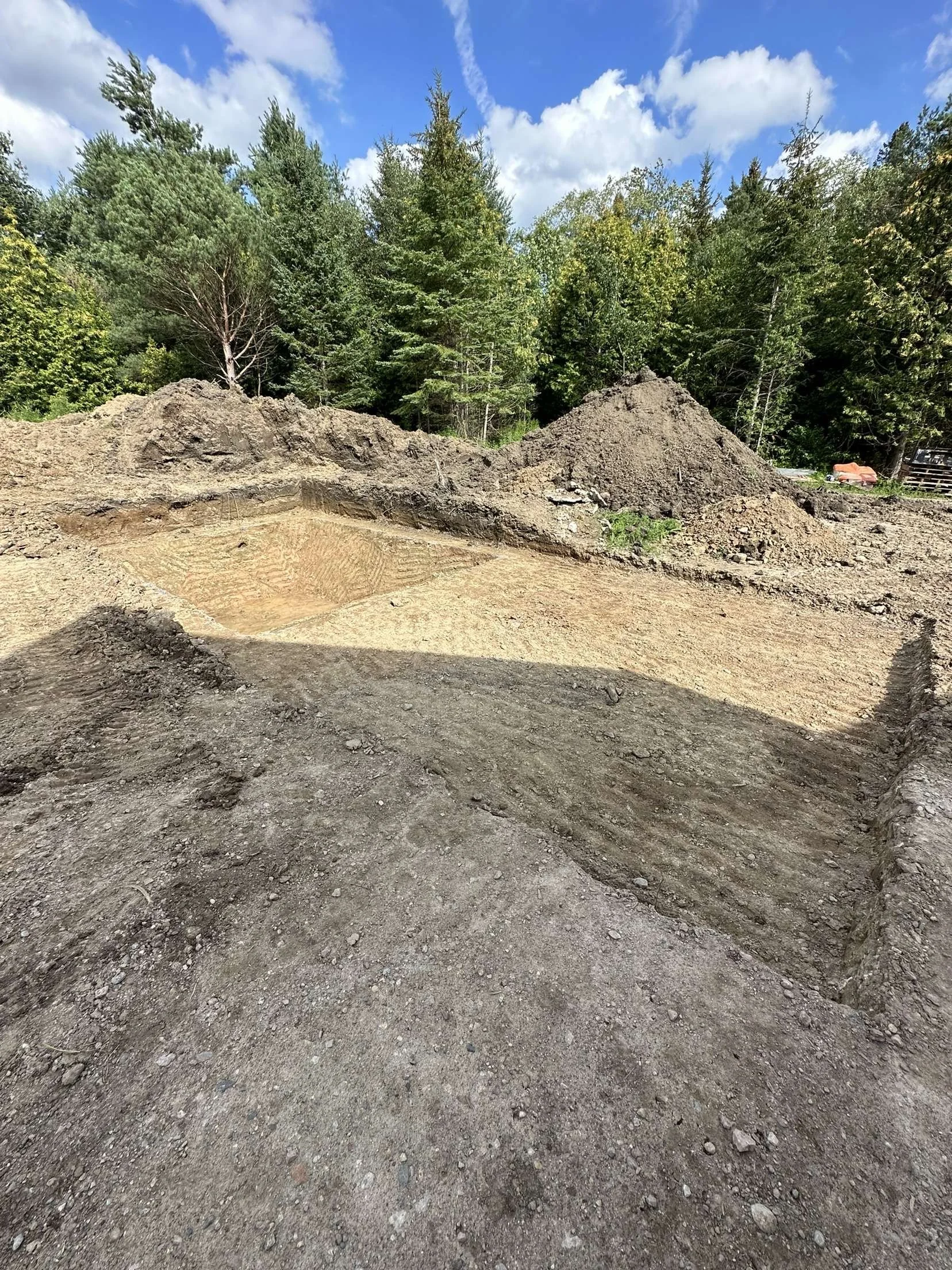 A construction site with excavated land, dirt mounds, and cleared earth, bordered by green trees under a partly cloudy blue sky.