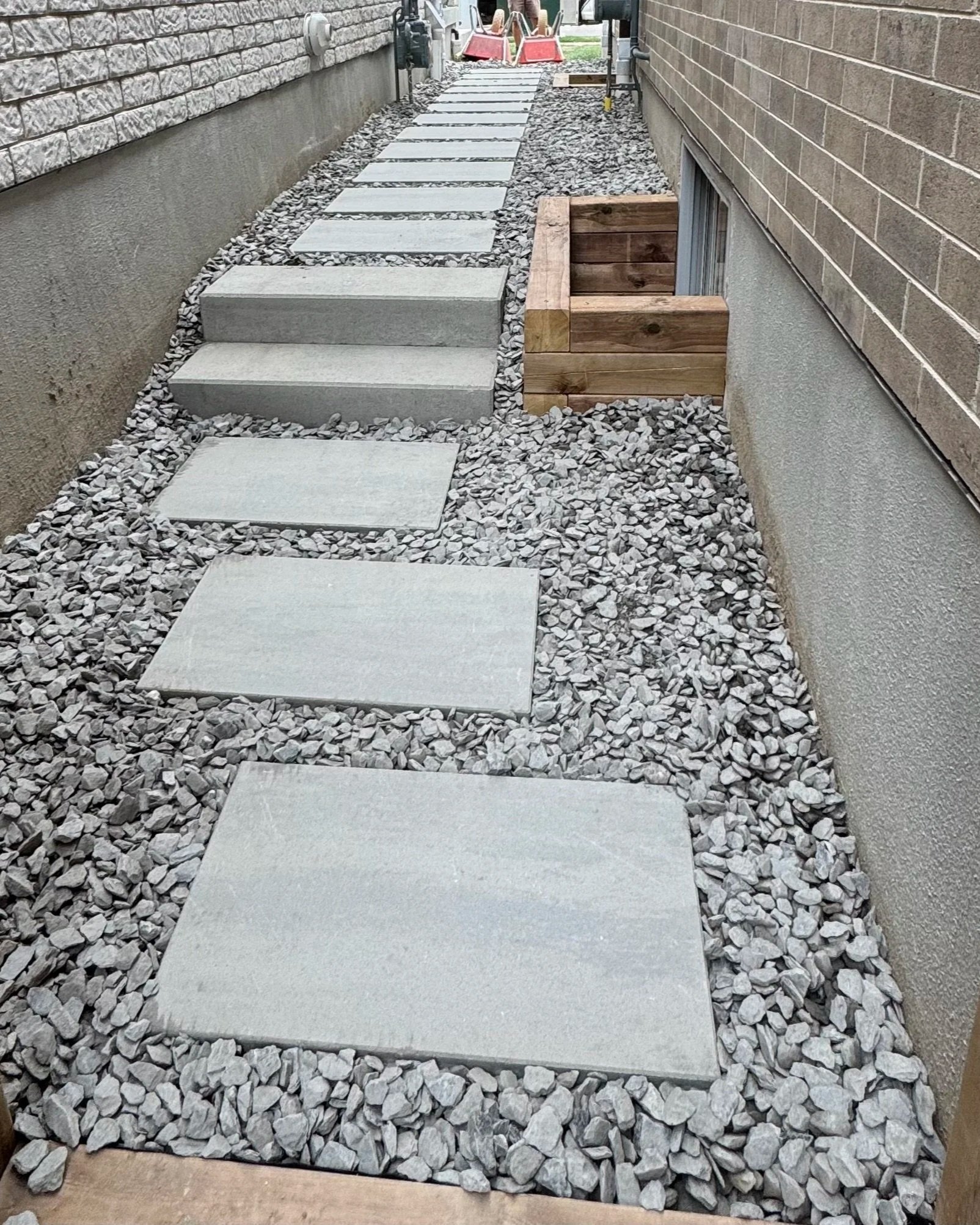 A narrow side yard with concrete stepping stones laid on gravel, bordered by brick walls on either side, with some wooden framing around a small window on the right wall.