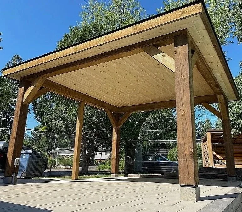 A wooden pavilion with a sloped roof, supported by wooden beams, situated on a paved area in an outdoor setting with trees around.
