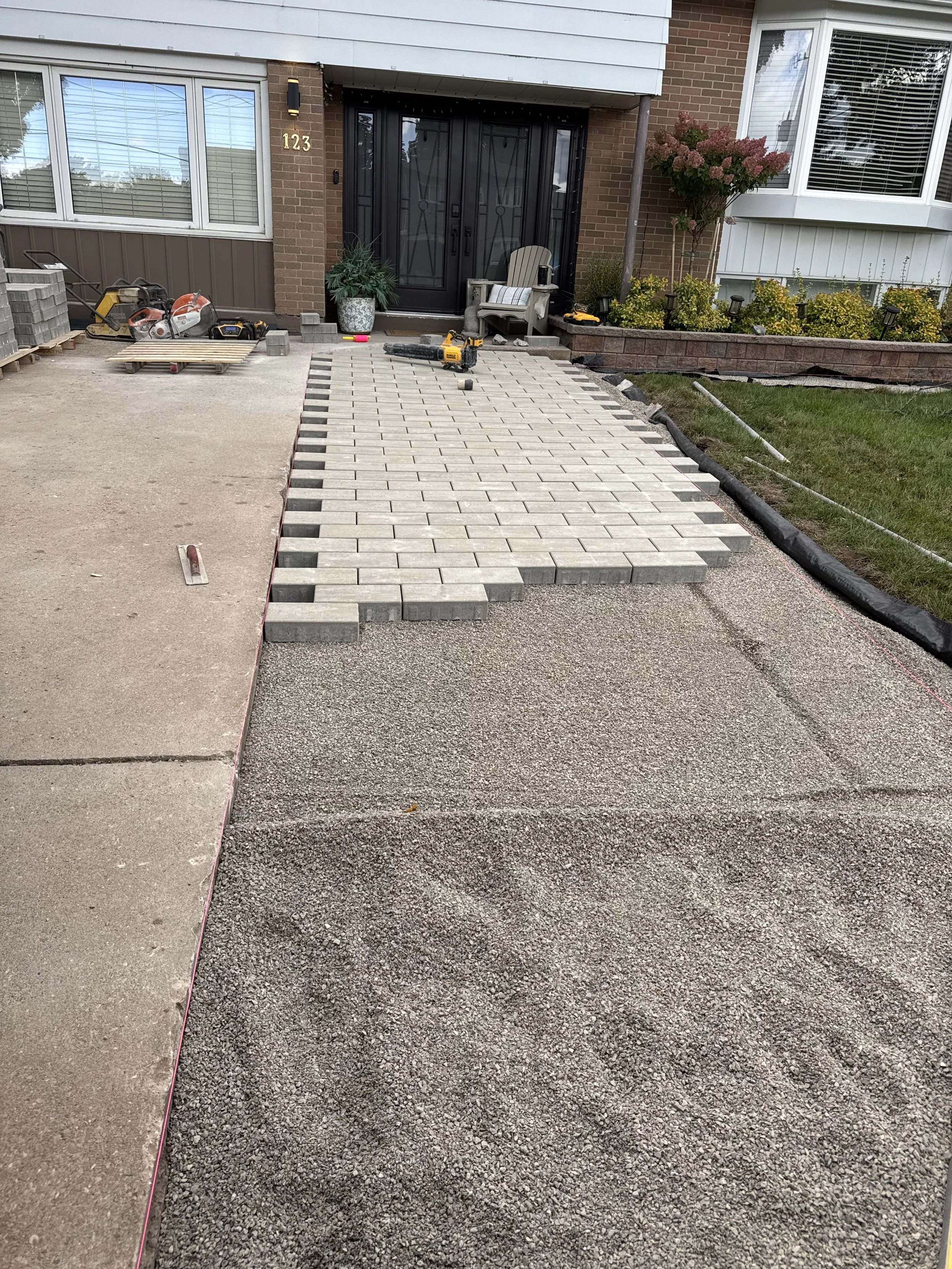 The image shows a house entrance with a partially completed brick or paver pathway. Construction tools and materials are present, and the area is in the process of being landscaped or renovated.