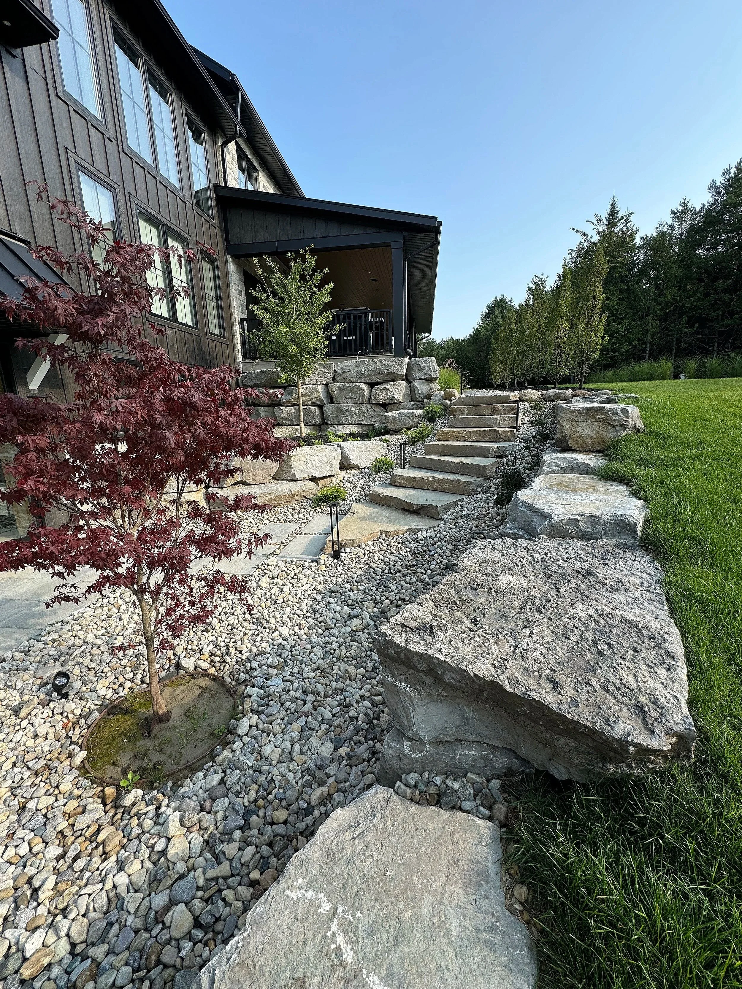 A modern house with a dark wooden exterior, large windows, and a covered porch. There are stone steps leading up to the porch, surrounded by landscaped rocks and plants, including a small tree with red leaves in the foreground. Green grass and taller trees are visible in the background under a clear blue sky.