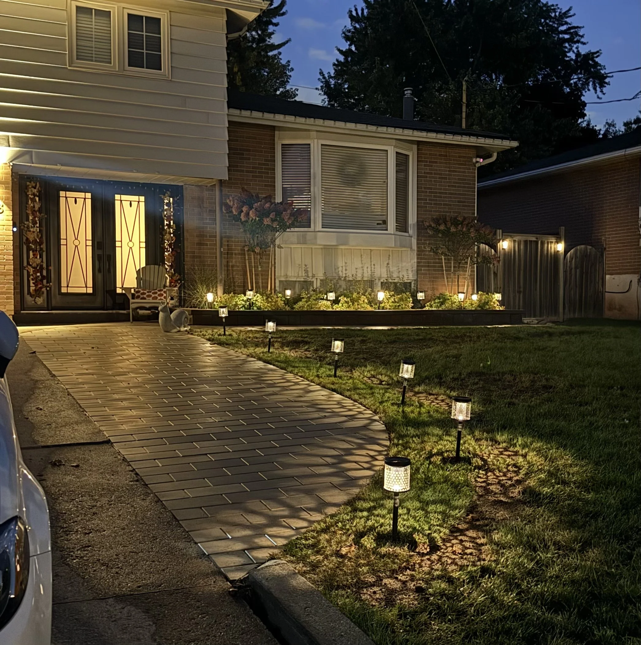 Exterior of a house at dusk with lights on inside, a pathway with small lantern lights, a lawn, and a gray cat sitting near the pathway. The house has brick and siding, multiple windows, and a front porch with doors decorated for the holidays.