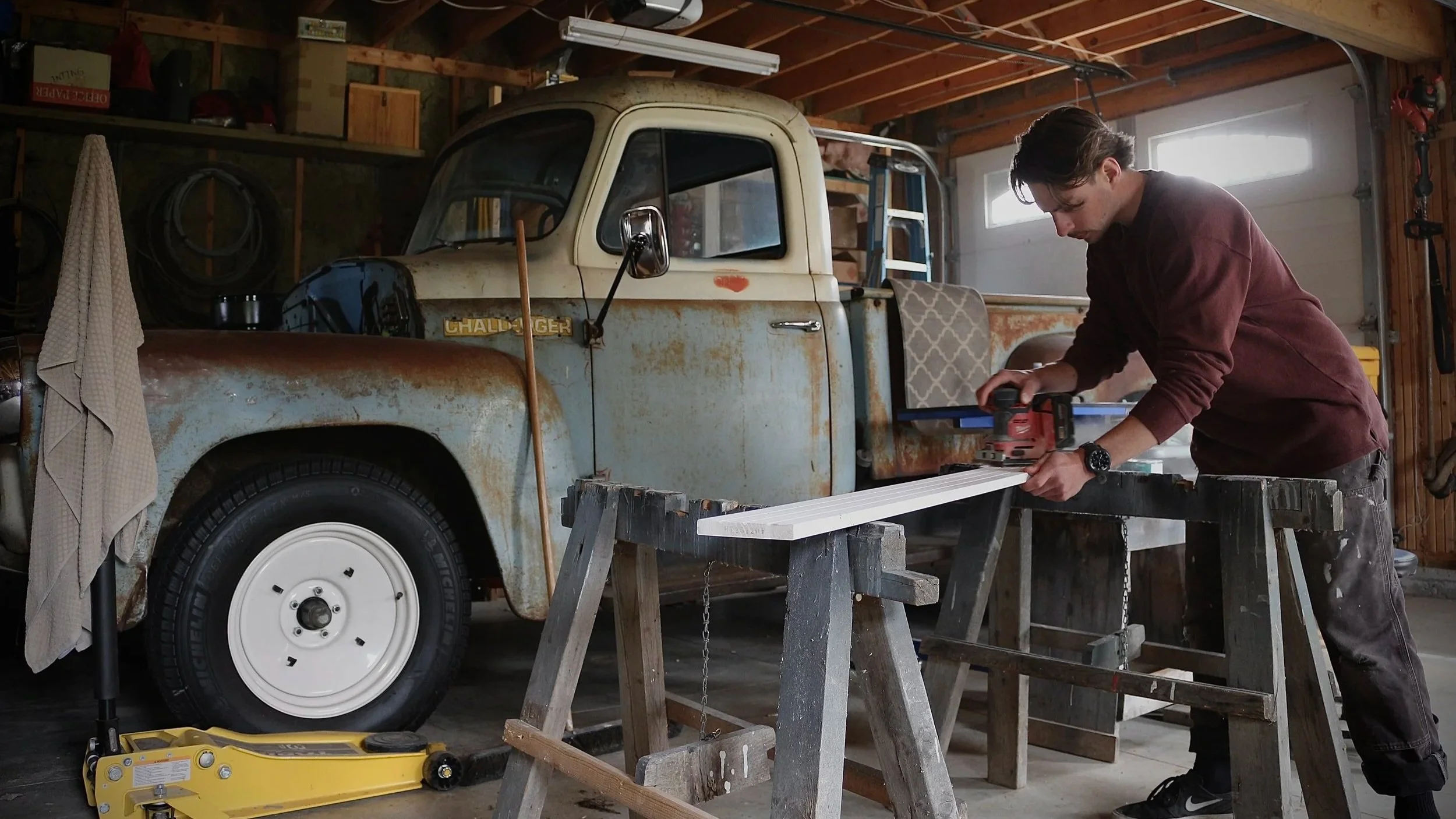 A young man working in a garage, using a power tool on a white wooden plank, with an old rusty vintage Dodge pickup truck nearby.