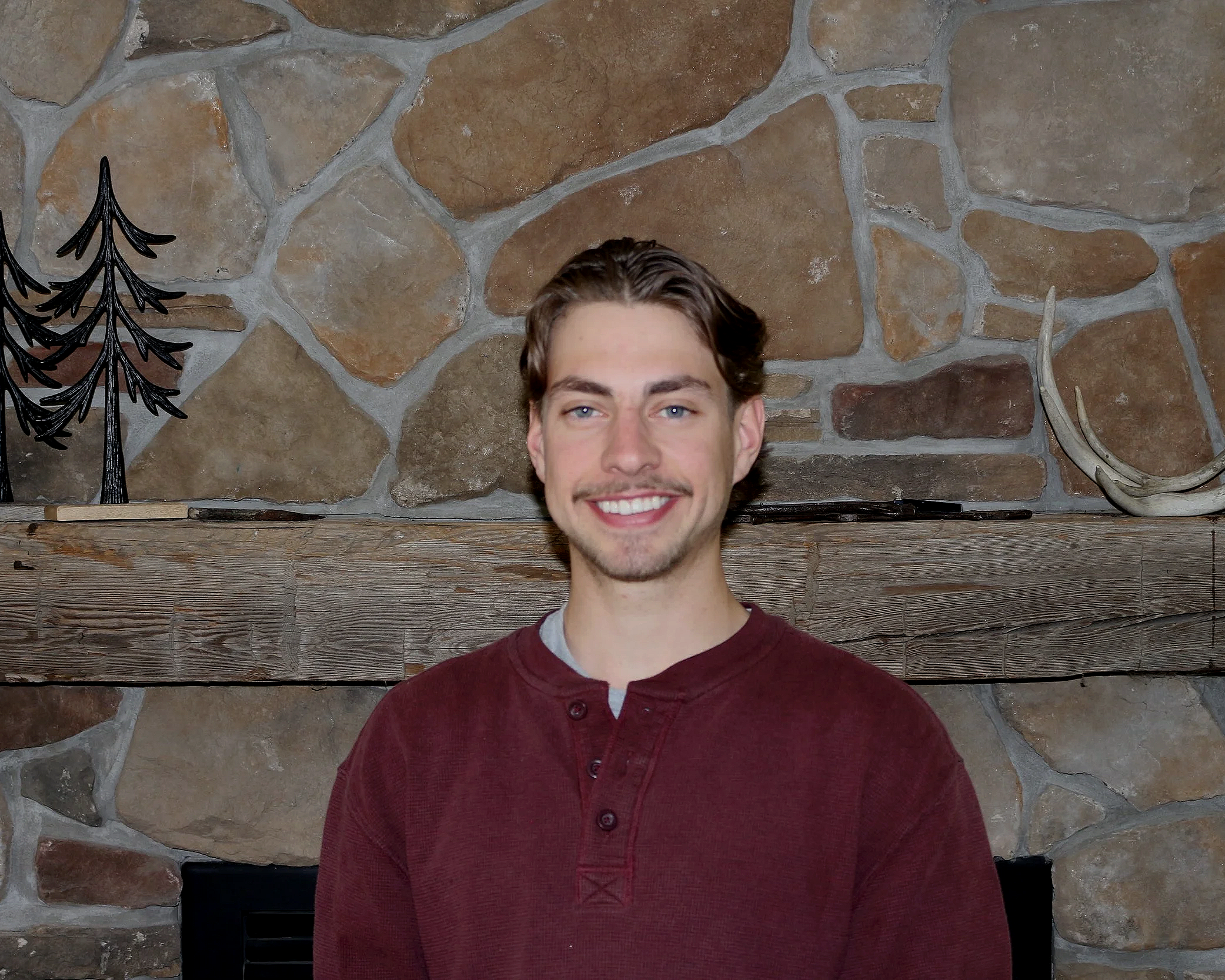 A young man with brown hair, blue eyes, and a mustache, smiling, is pictured with a background of a stone wall and wooden shelf. He is wearing a maroon button-up shirt.