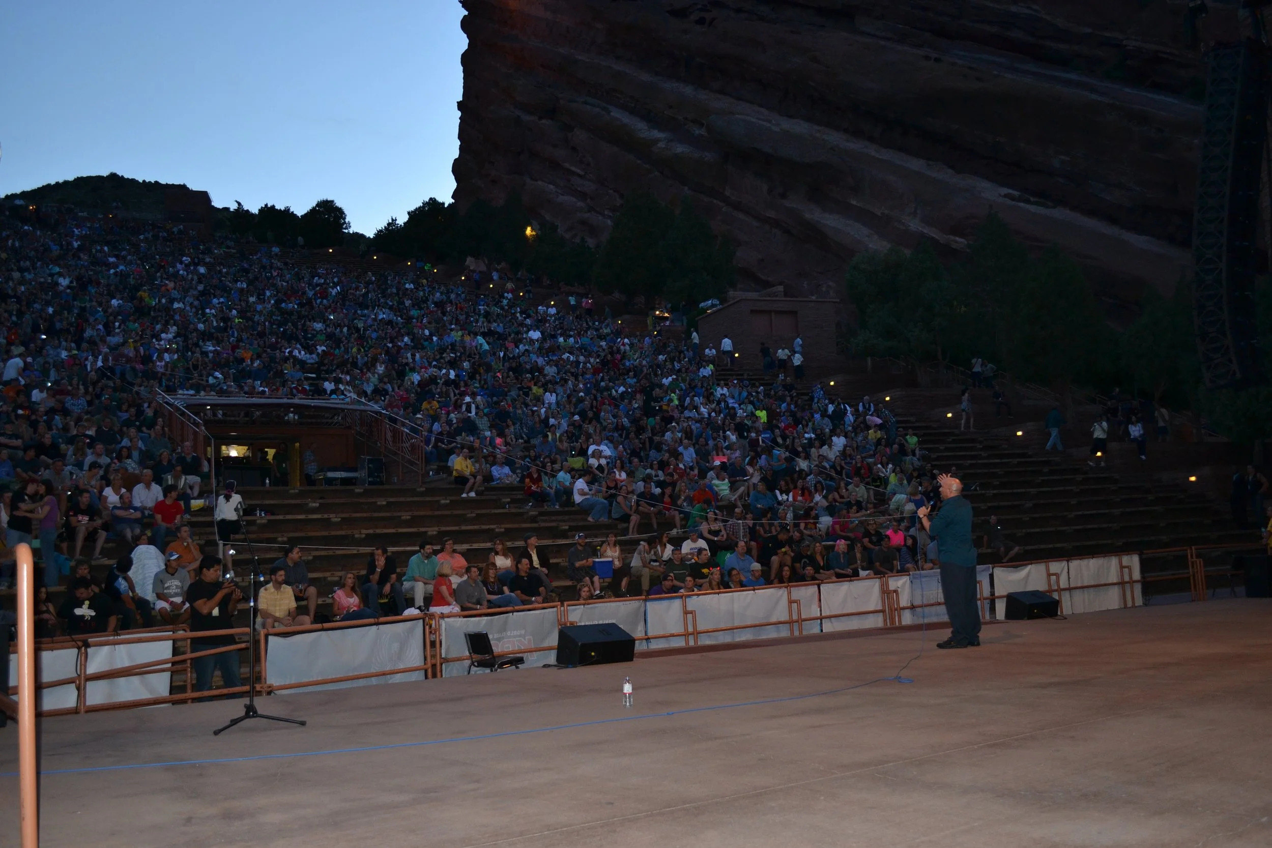 Vic Alejandro performing stand-up comedy at Red Rocks outdoor Amphitheatre in Colorado filled with a large crowd of people seated on tiered stone benches under a cliff face during twilight.