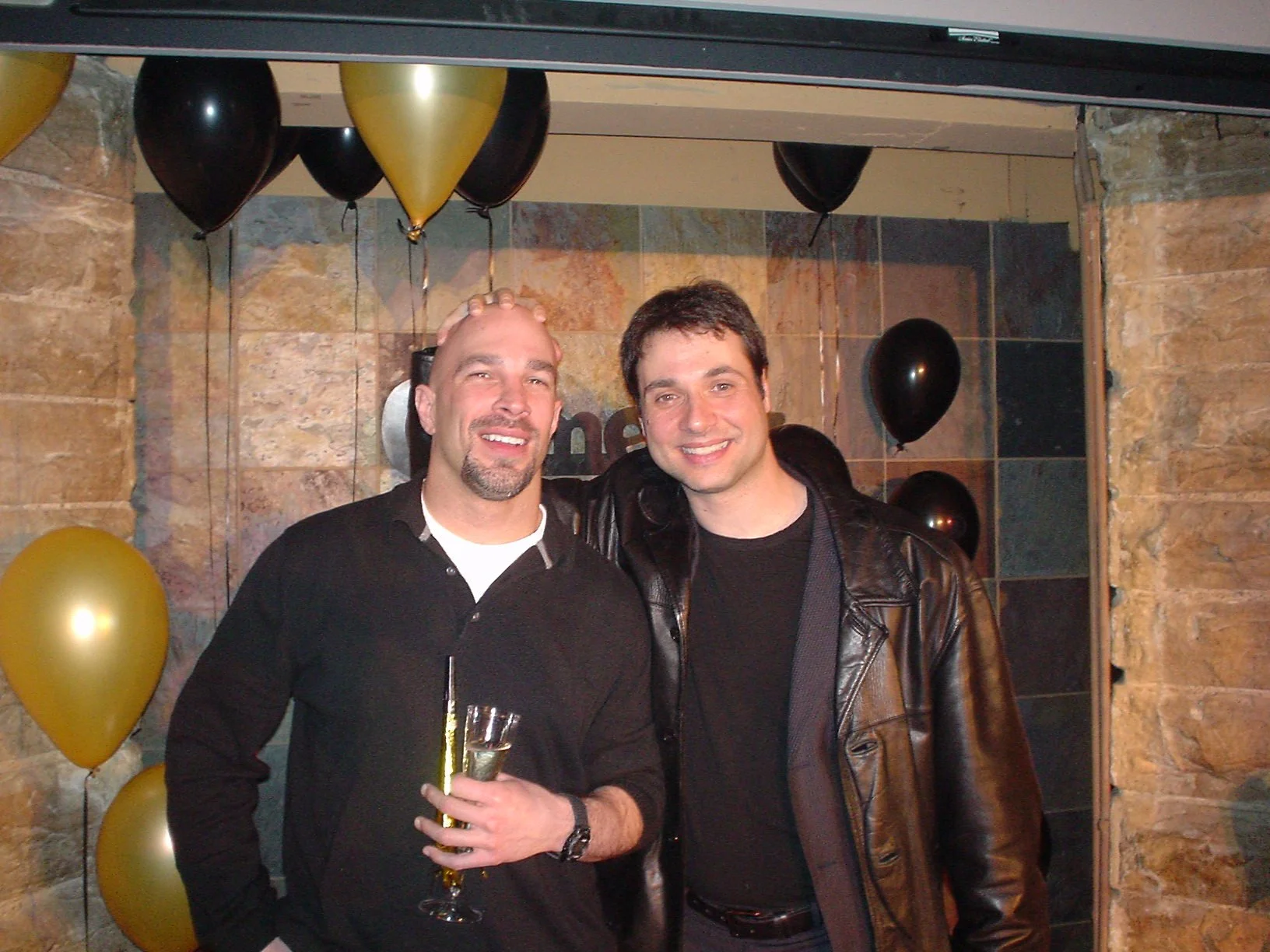 Vic Alejandro with comedian Adam Ferrara on stage at Comedy Works downtown. Two men standing together, smiling at a New Years celebration with black and gold balloons in the background, holding a glass of champagne. 