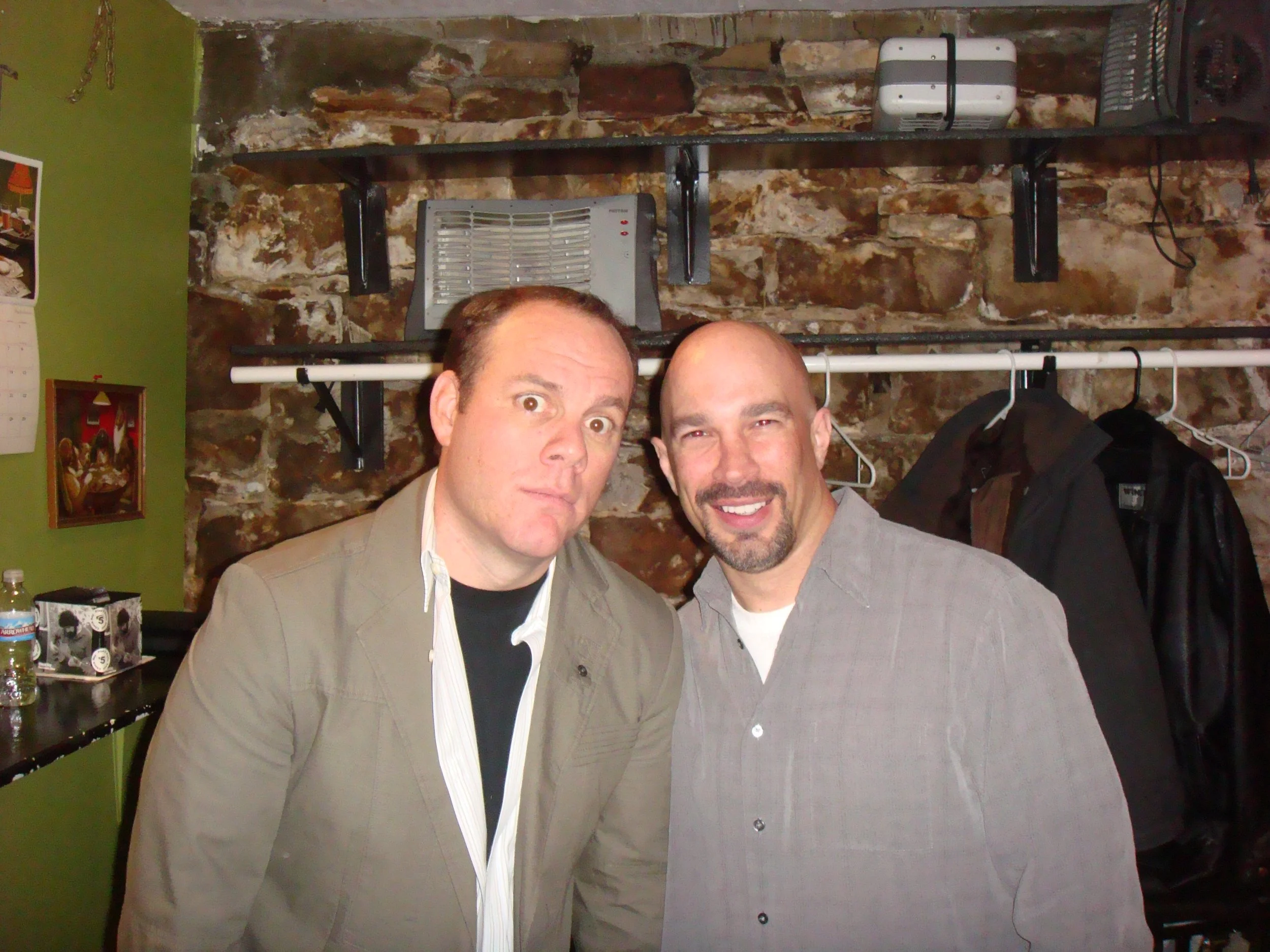 Vic Alejandro with comedian Tom Papa in the green room of Comedy Works in downtown Denver, Colorado.  Posing for a photo in front of a brick wall . They are both smiling, one with a surprised expression and the other smiling broadly.