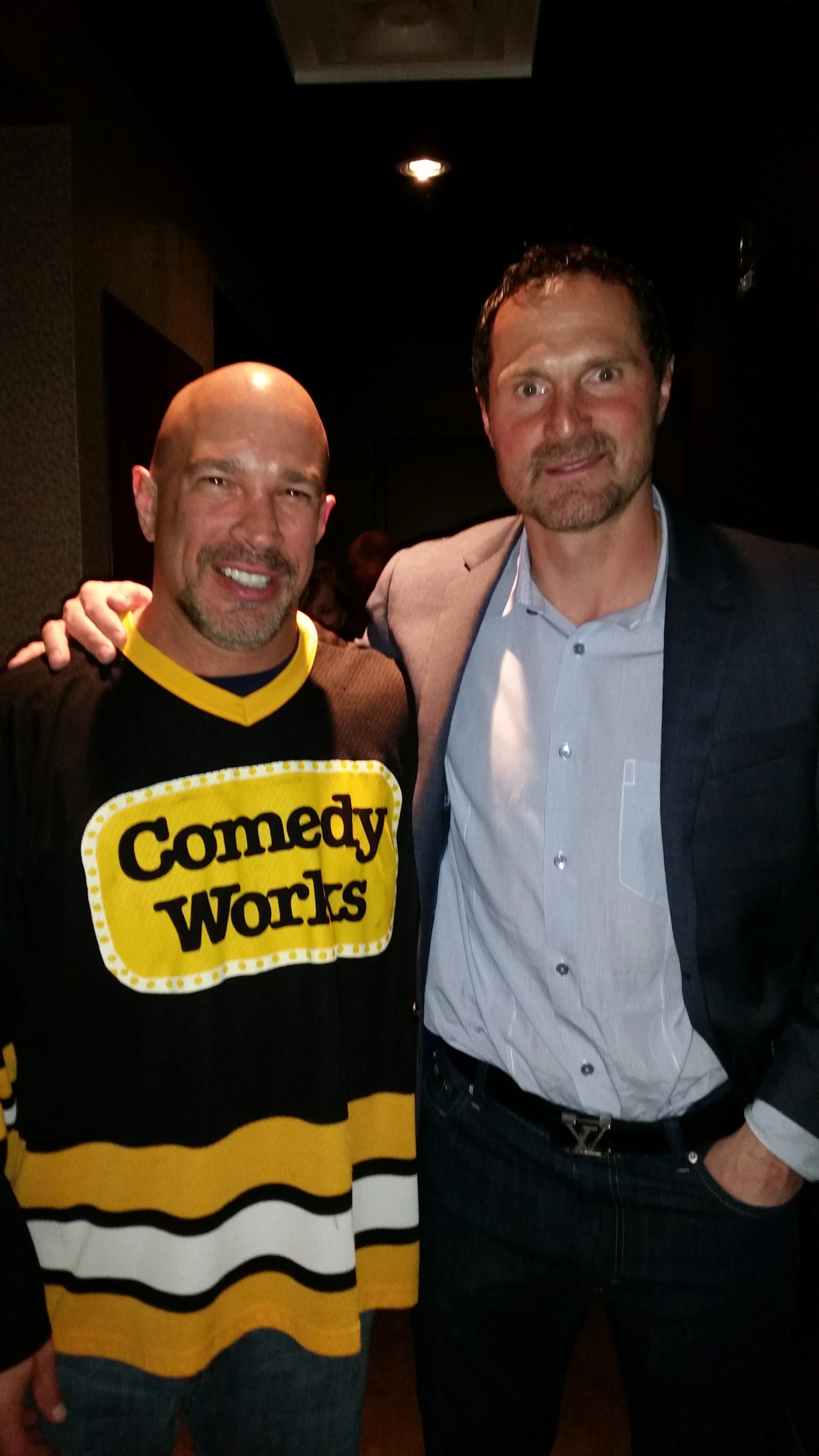 Vic Alejandro with former NHL and Colorado Avalanche great Milan Hejduk. Two men standing close together, Vic wearing a black and yellow 'Comedy Works' hockey jersey, and Milan in a gray blazer and light blue shirt. 