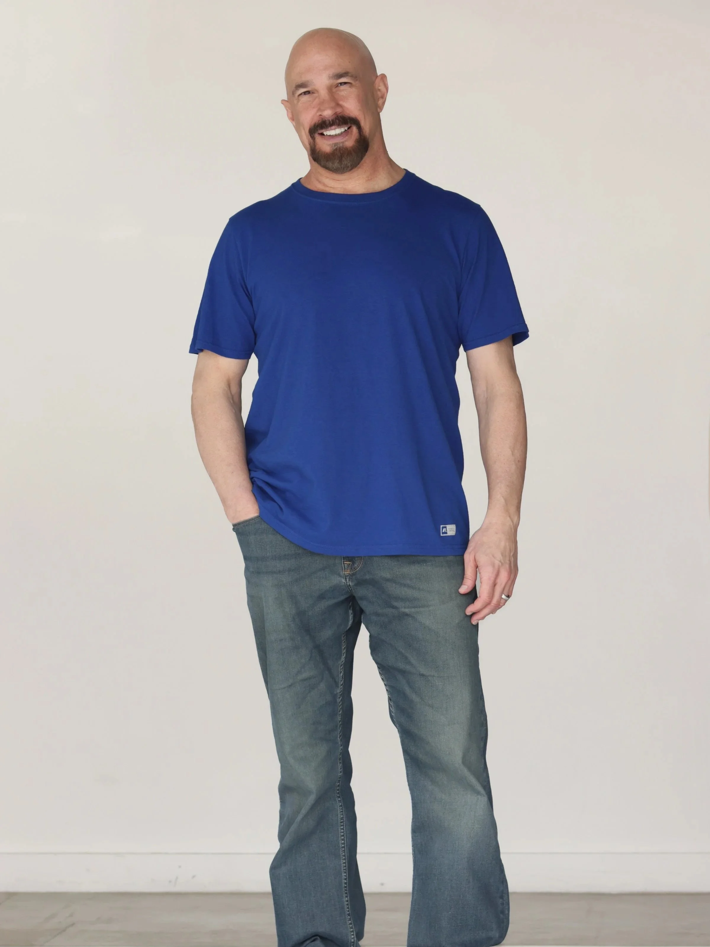 Vic Alejandro in a full length body shot smiling with a goatee wearing a blue t-shirt and jeans standing against a plain white wall.