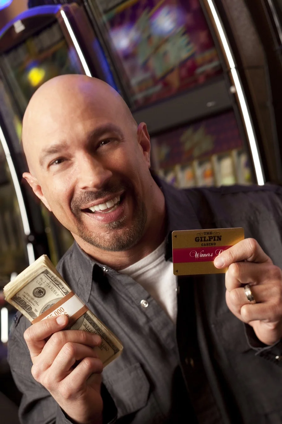 Vic Alejandro holding a stack of hundred-dollar bills in one hand and a casino rewards card in the other, smiling in front of a slot machine at a casino.