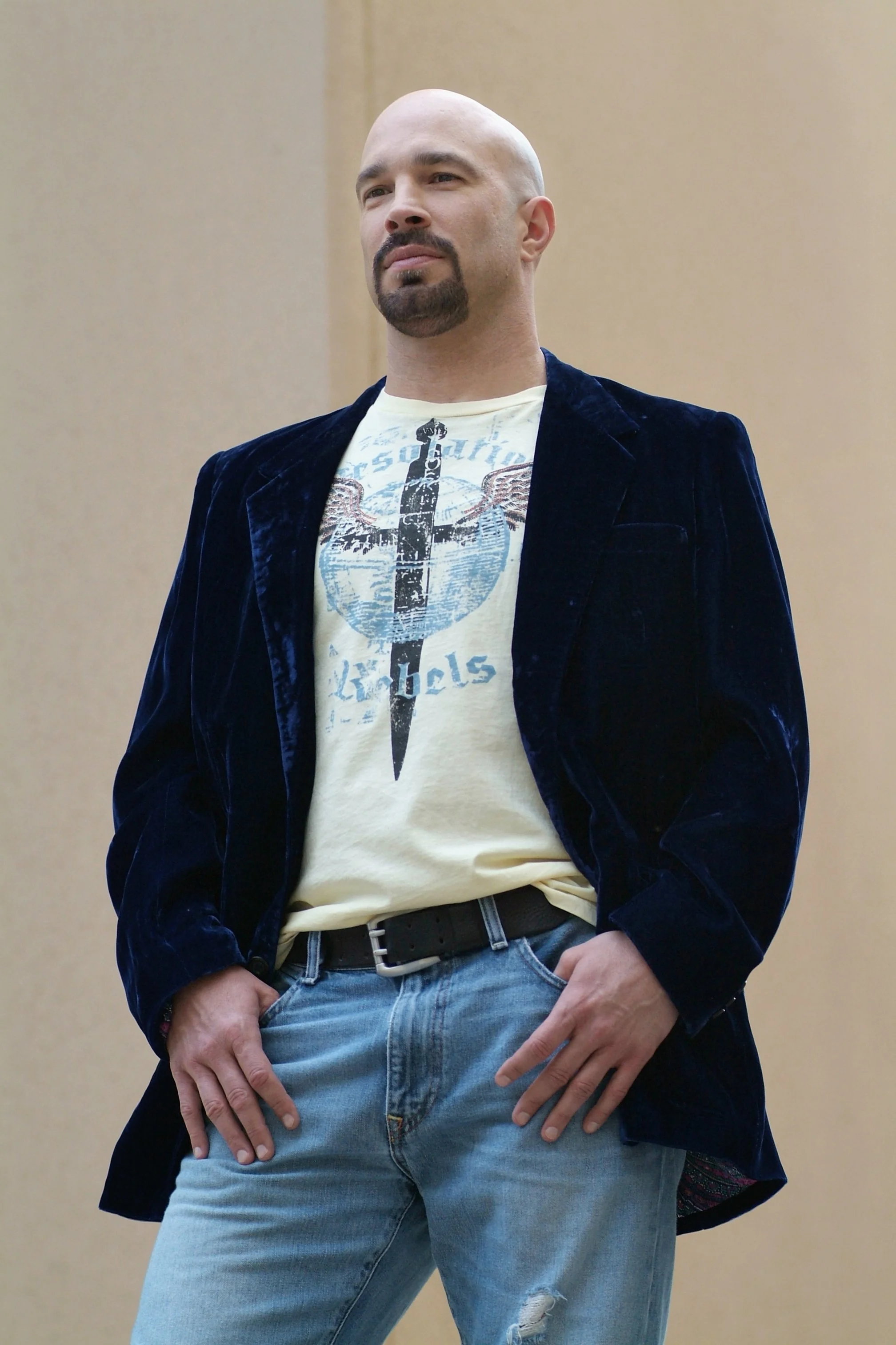 Vic Alejandro with a shaved head, goatee, and mustache wearing a navy blue velvet blazer, a graphic T-shirt with a tattoo design, and light blue ripped jeans with a black belt, standing with hands in his pockets against a beige wall.