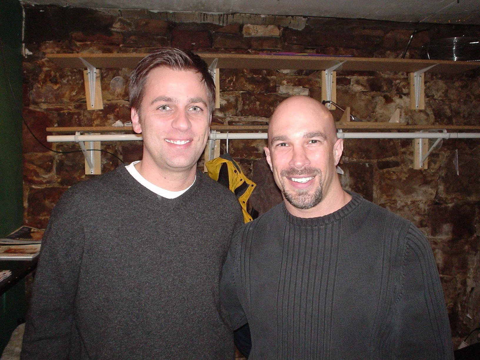 Vic Alejandro with comedian John Heffron in the green room of Comedy Works in downtown Denver, Colorado. Two men standing closely together, smiling for the camera in front of a brick wall with shelves. 
