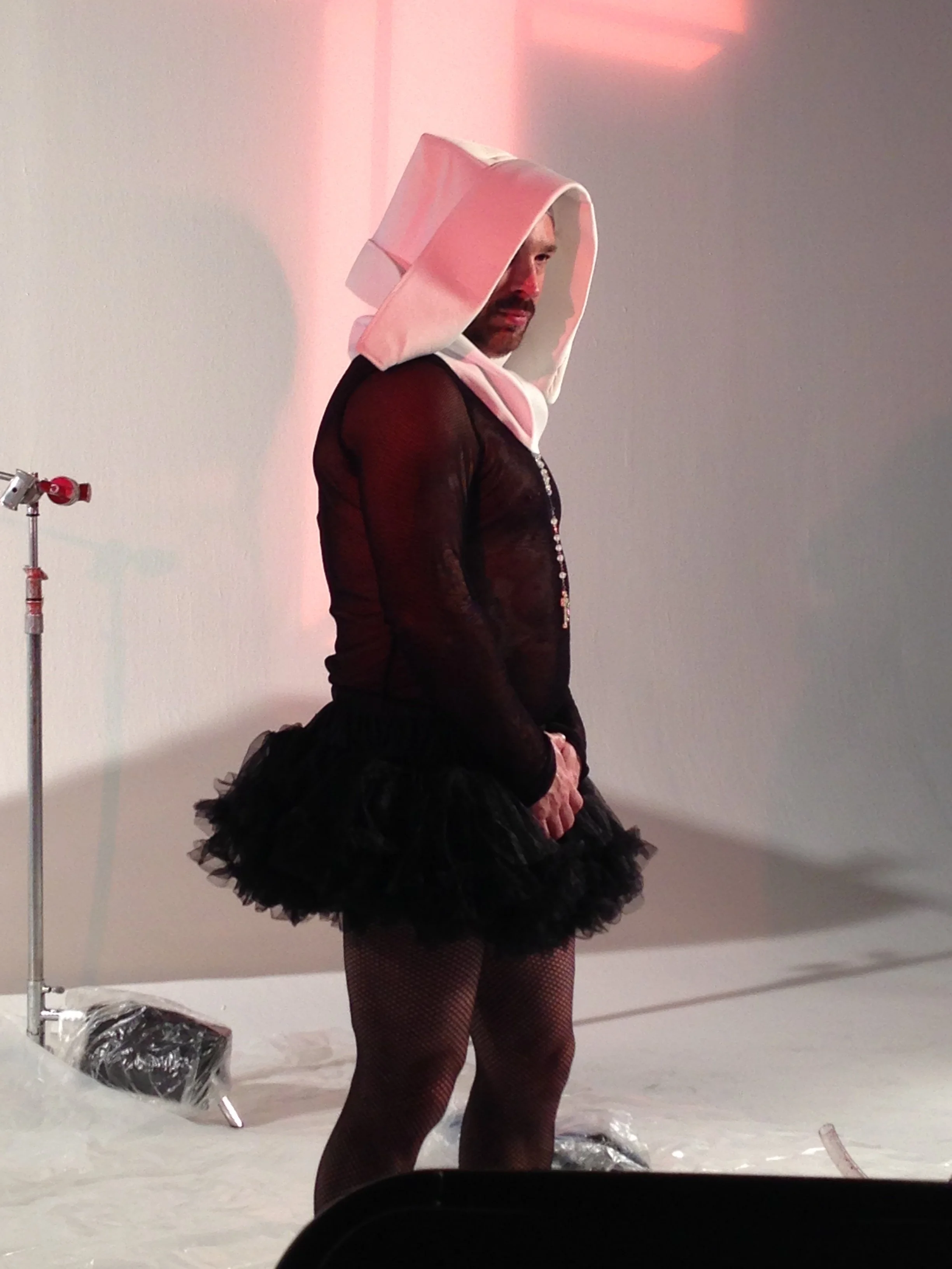 Vic Alejandro on the set of "Directors on Directing" dressed as a nun in a white habit and black tutu skirt standing in a studio with a large illuminated cross on the wall behind them. He is just standing getting ready.