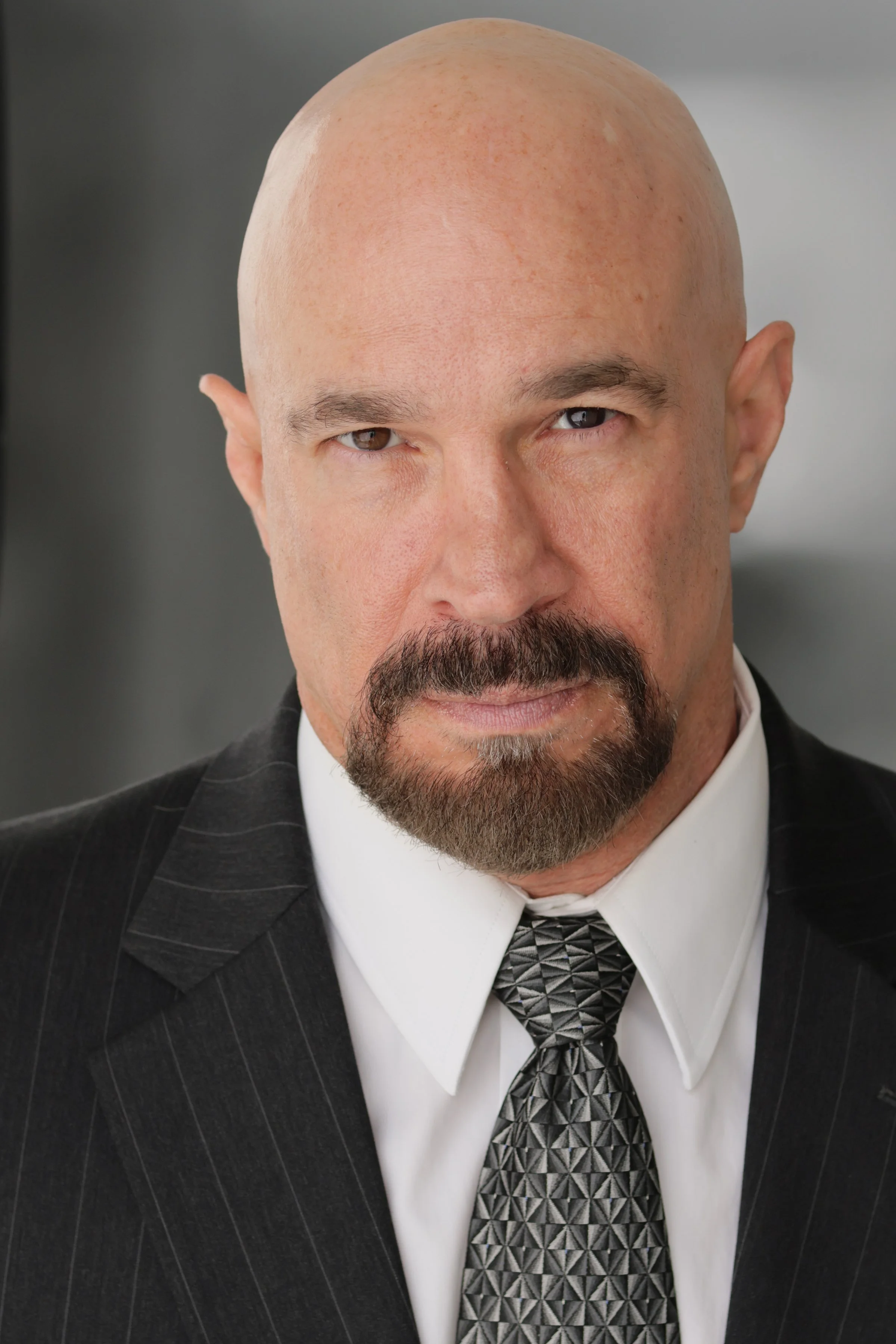 Vic Alejandro with a bald head, brown eyes, a goatee, and mustache dressed in a pinstripe suit, white shirt, and patterned tie.