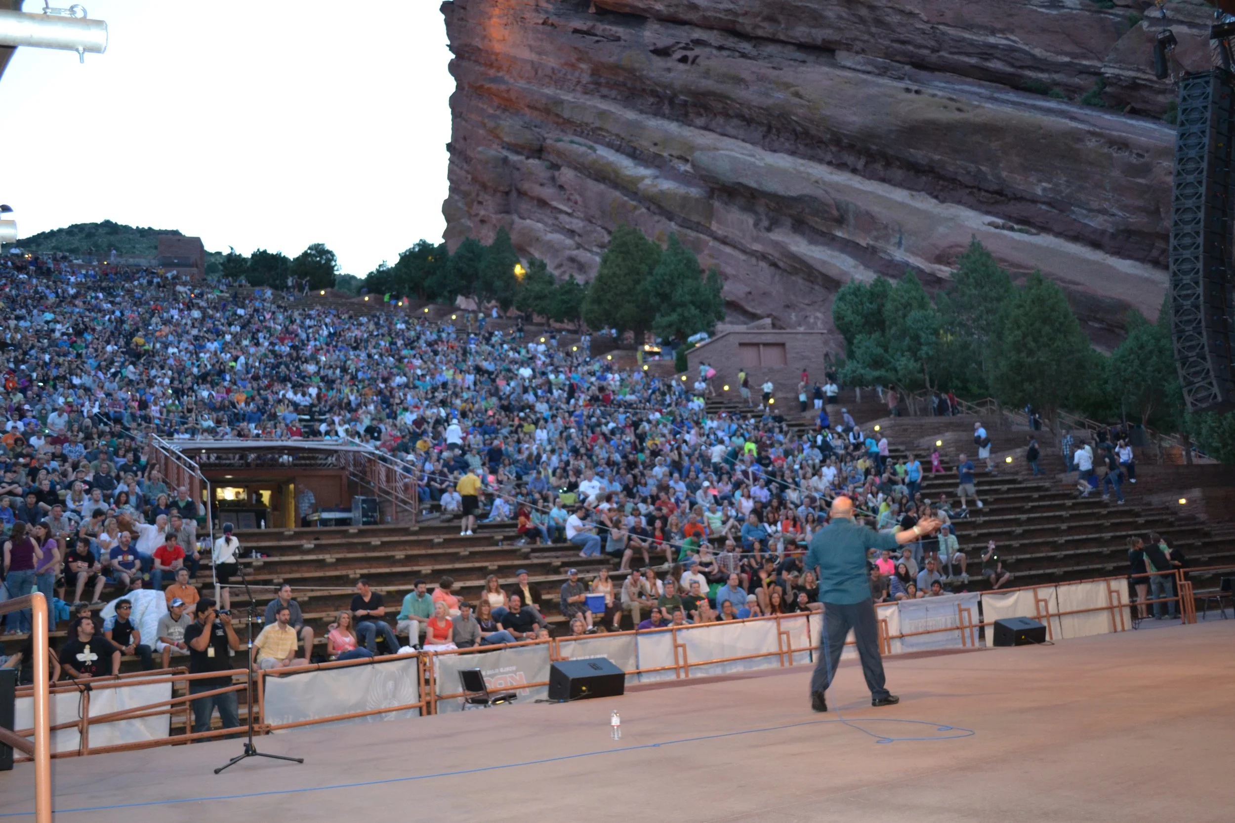 Vic Alejandro performing stand-up comedy at Red Rocks outdoor Amphitheatre in Colorado filled with a large crowd of people seated on tiered stone benches under a cliff face.