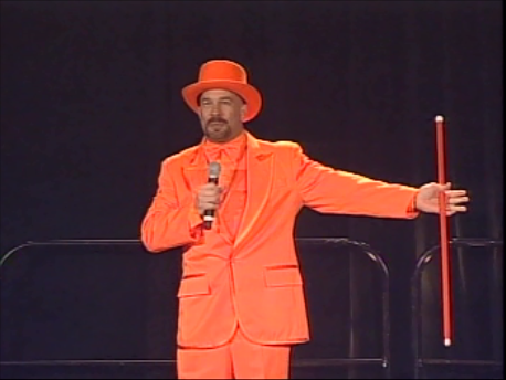 Vic Alejandro in an orange tuxedo, orange hat, and holding an orange cane standing on a stage with a black backdrop, speaking into a microphone at the PMI Conference in Denver, Colorado.
