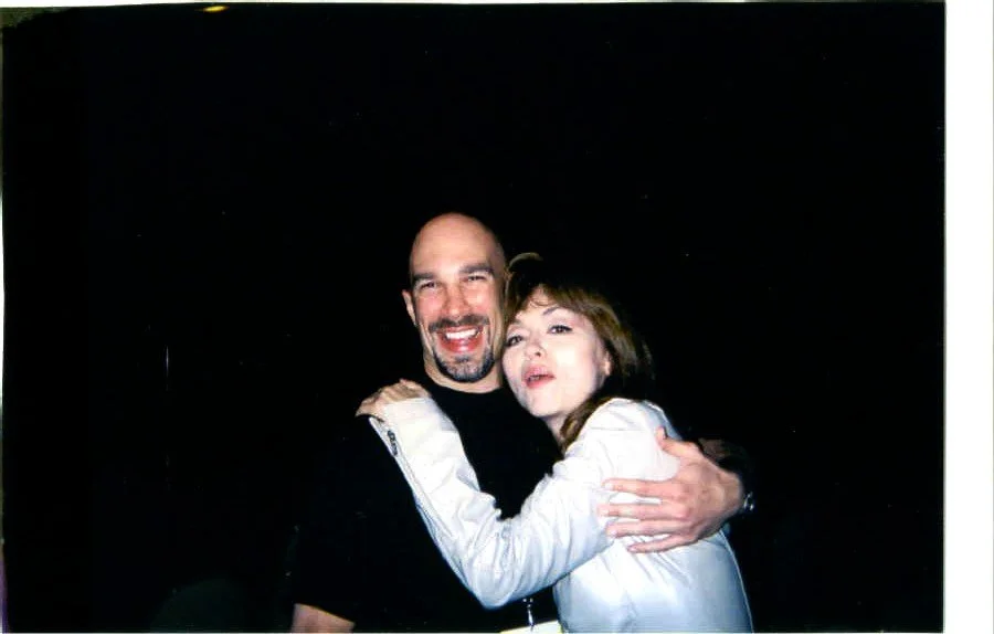Vic Alejandro with comedian Judy Tenuta (RIP) at a comedy festival in Las Vegas. A man and woman l hugging each other and smiling, against a dark background.