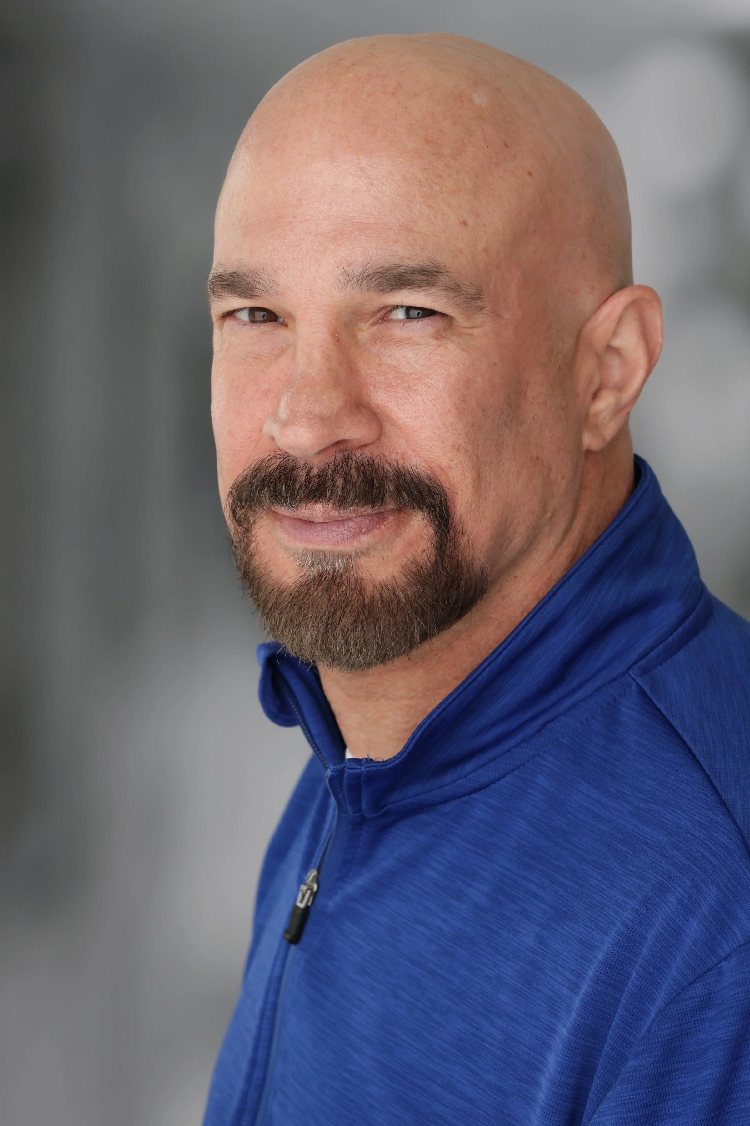 Vic Alejandro with a beard and mustache, wearing a blue jacket, smiling slightly, with a blurred gray background.