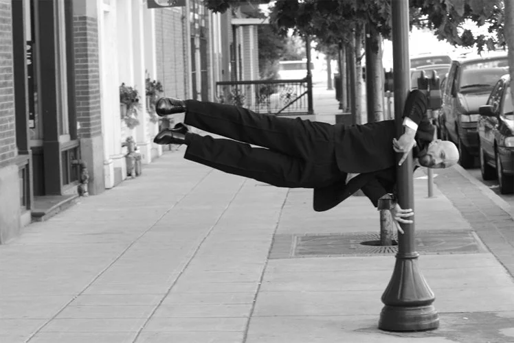 Vic Alejandro in a suit is balancing horizontally on a lamp post with his body stretched out, holding onto a street lamp post on a sidewalk.